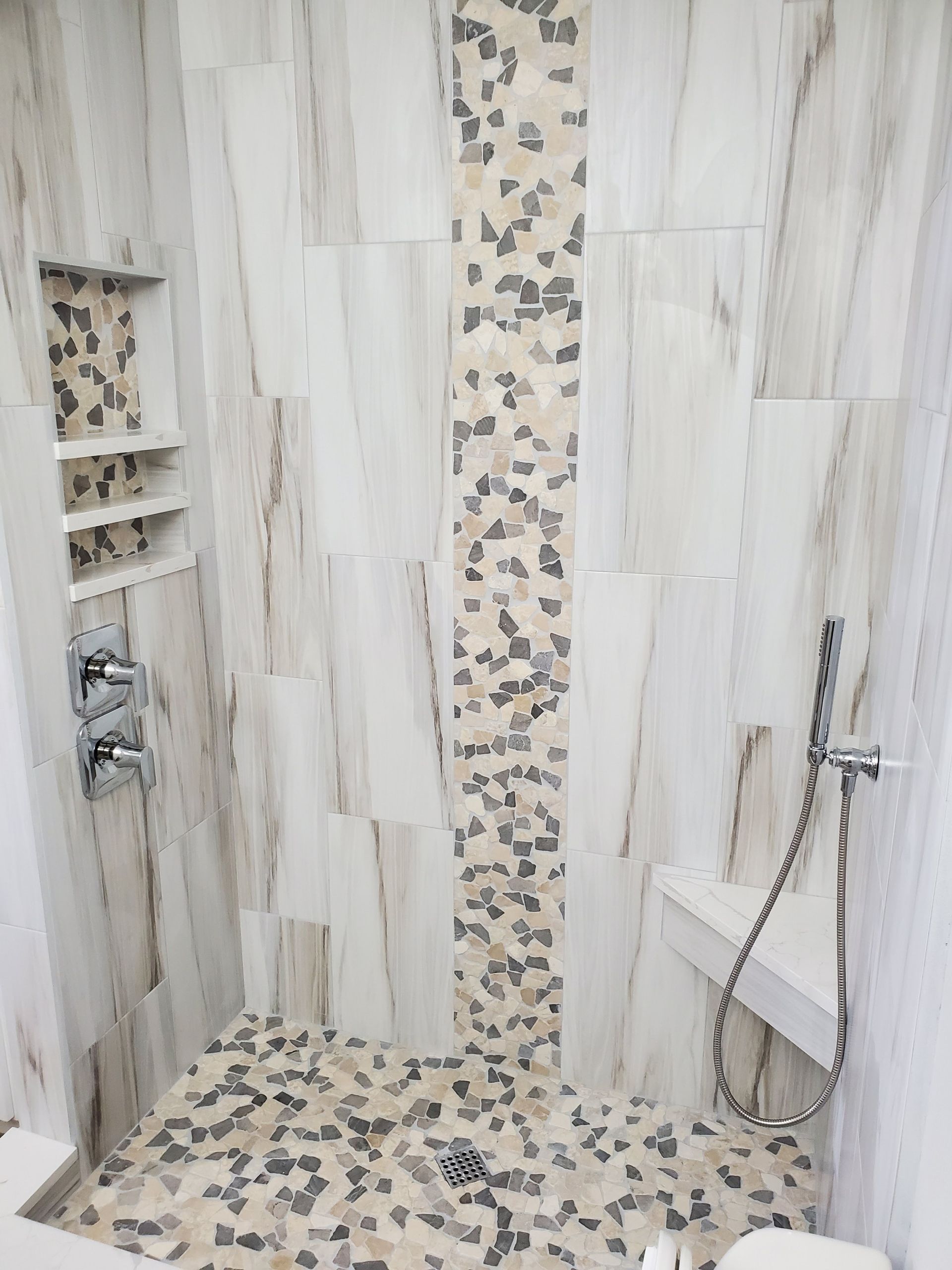 A walk-in shower with vertical light marble tiles, a central pebble stone accent strip, matching pebble floor, and bench.