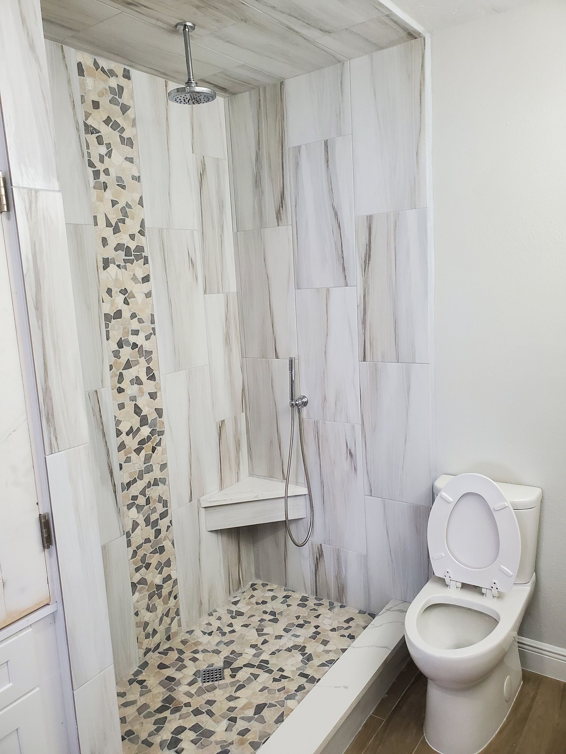 A modern walk-in shower with white and grey marble tiles, a pebble-stone floor, and a built-in seat, next to a toilet.