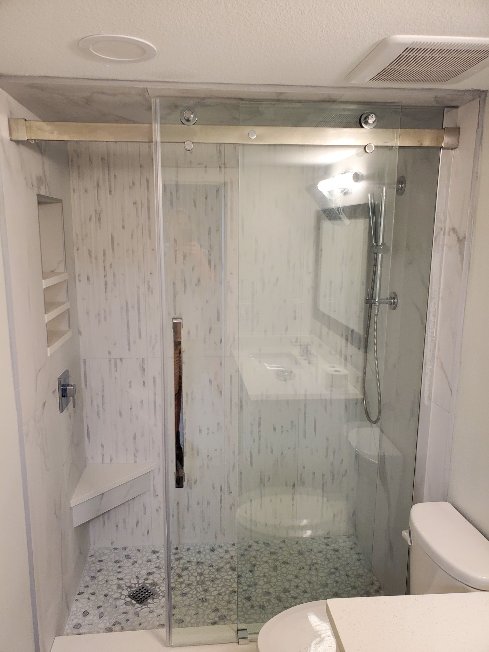 A walk-in shower with a glass sliding door, marble-patterned tile walls, a corner bench, and pebble-style floor tile.