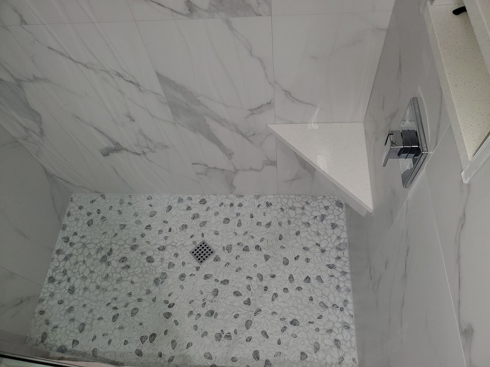 High-angle view of a walk-in shower with white marble-look wall tiles, a pebble-stone floor, and a triangular corner shelf.