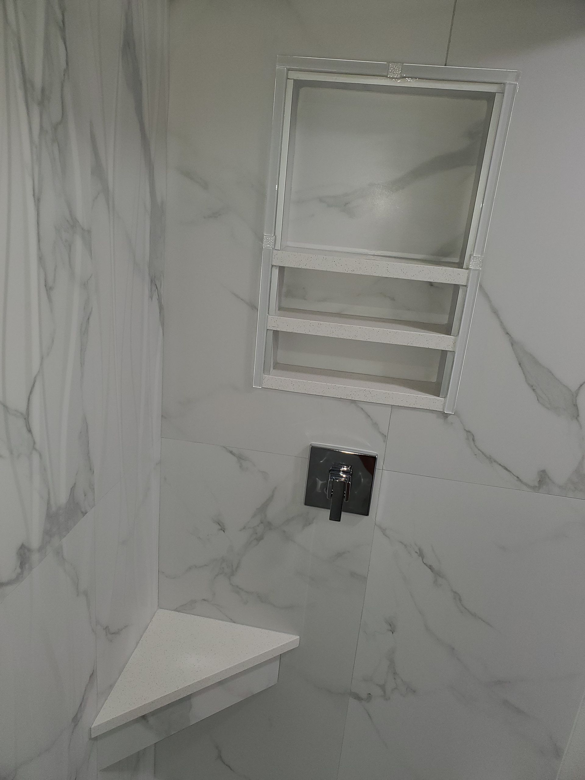 A white marble-patterned shower wall with a built-in recessed shelf and a corner shelf near a faucet handle.