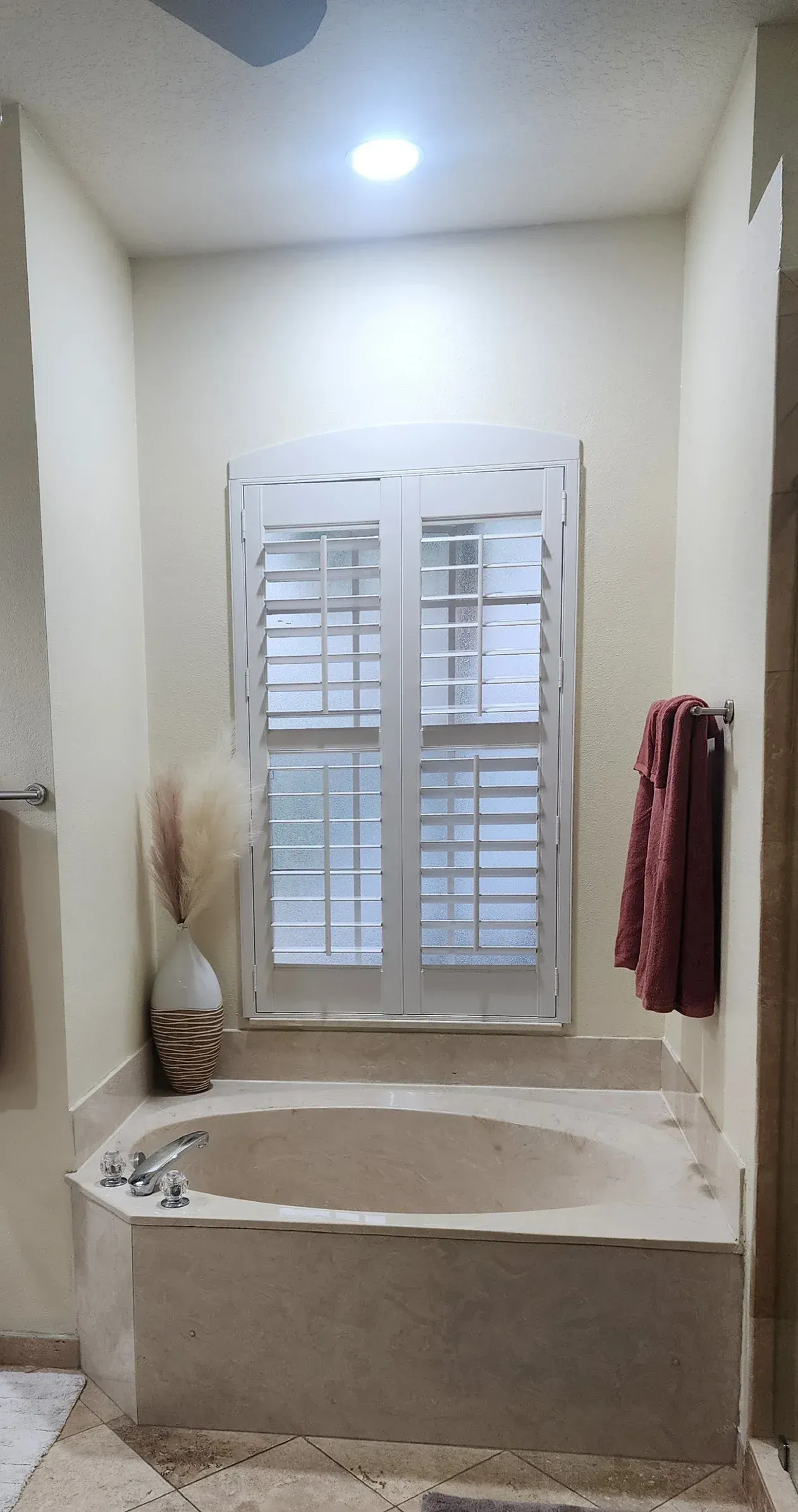 Bathroom with a built-in bathtub, a window with shutters, and towel rack. Beige walls and light fixture.