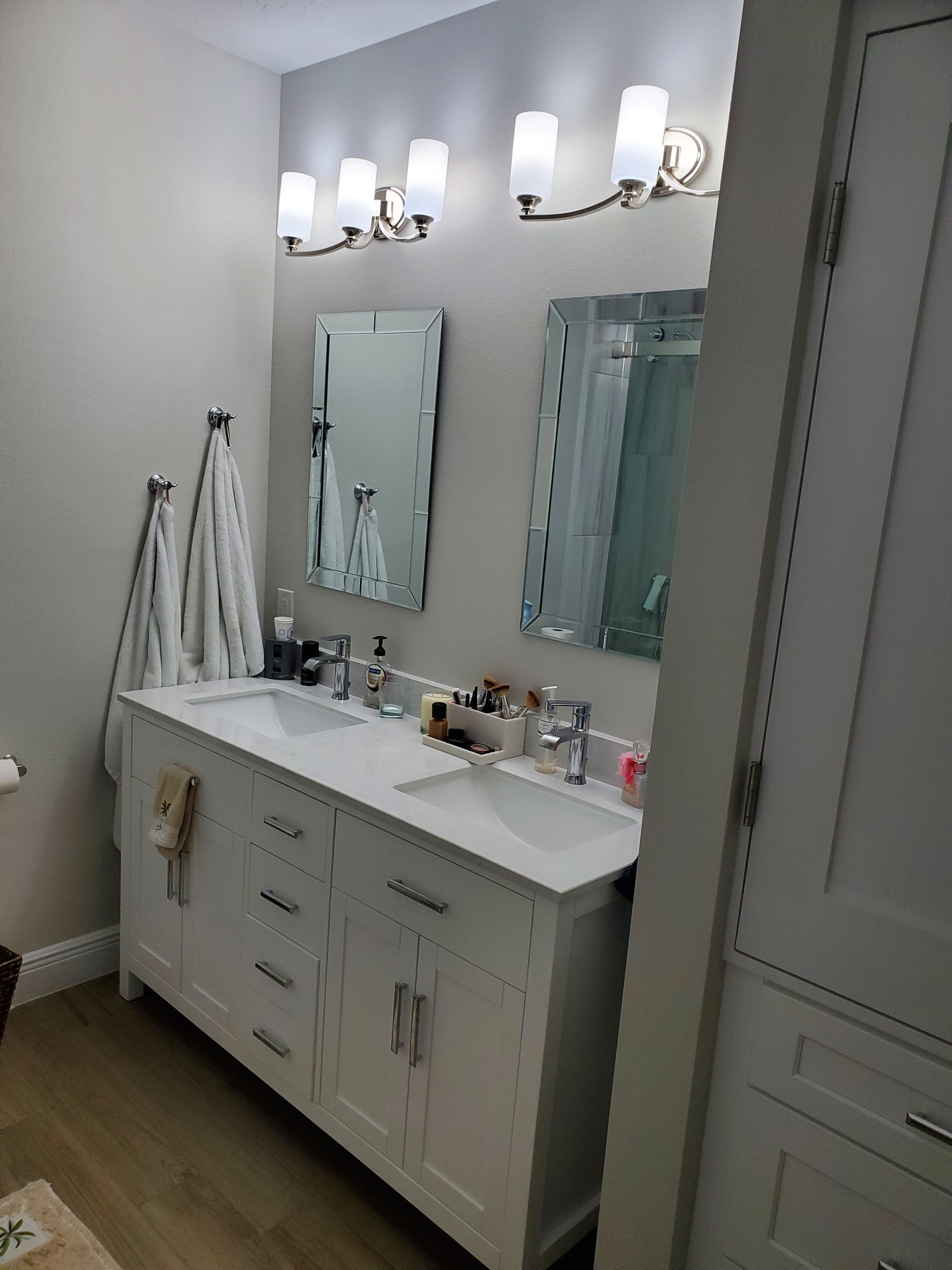 A white double-sink bathroom vanity with two mirrors, silver fixtures, and overhead lighting in a neutral-toned bathroom.
