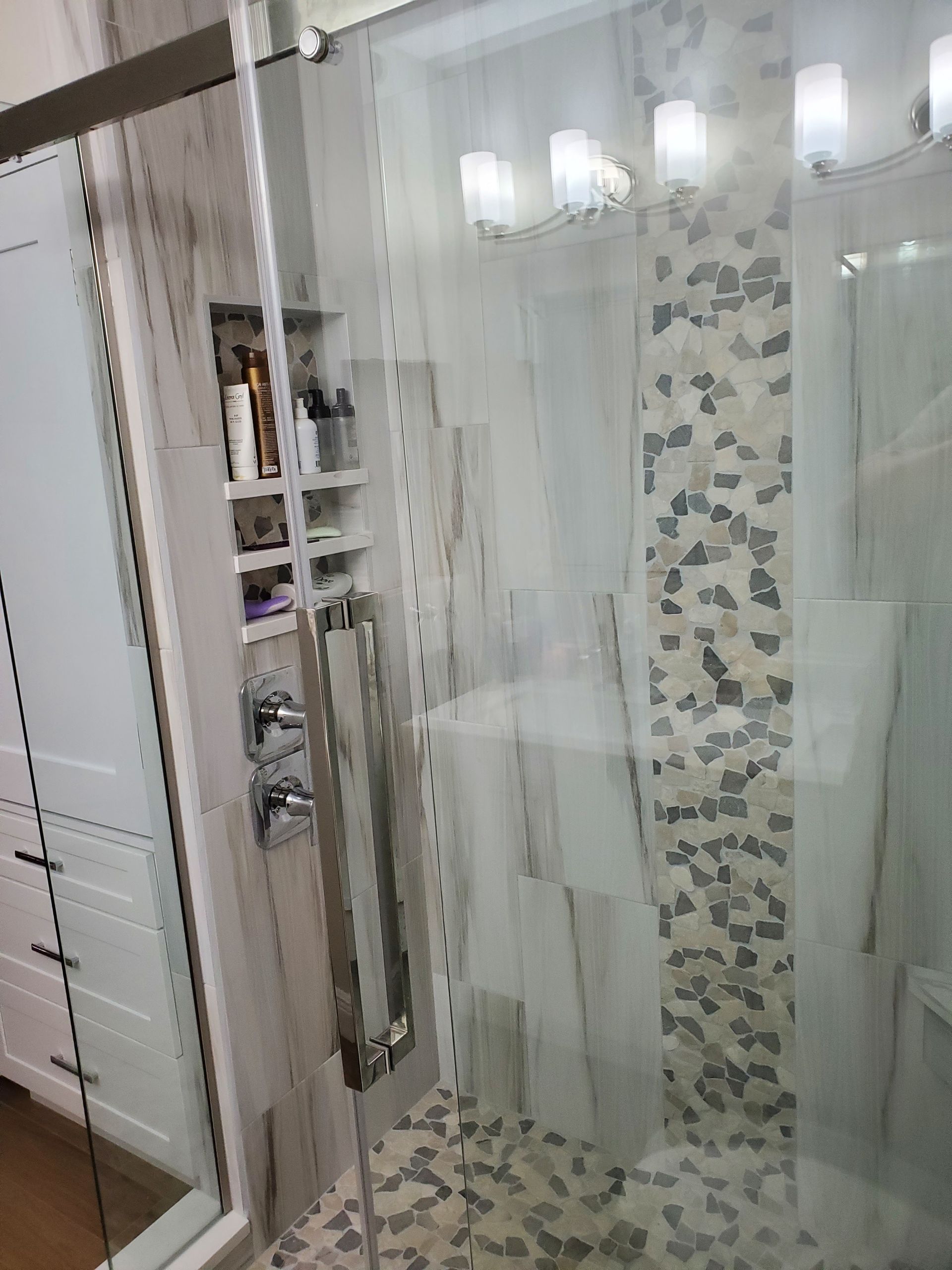 A modern glass shower with a tiled niche containing bottles, a stone-patterned accent wall, and a mosaic tiled floor.