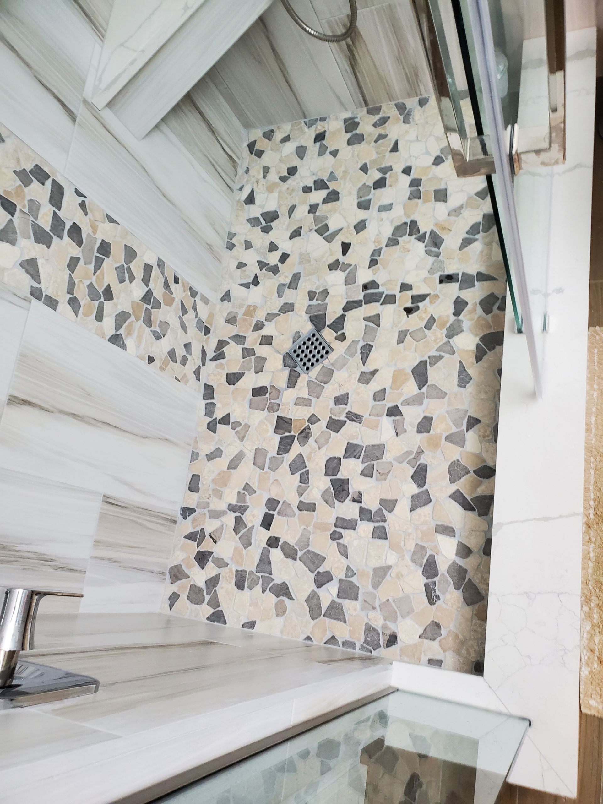 A modern shower with large, light wood-look wall tiles and a multi-colored stone mosaic tile floor featuring a drain.