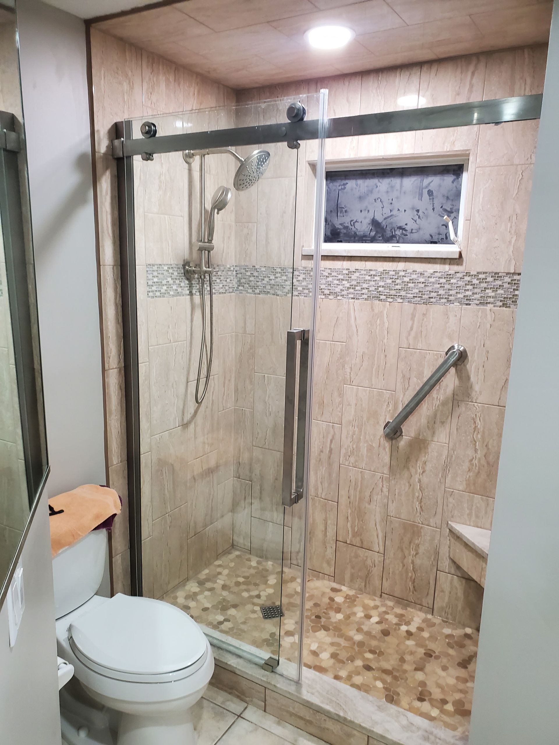 A tiled walk-in shower with a glass door, a handheld showerhead, a built-in seat, and a grab bar next to a toilet.