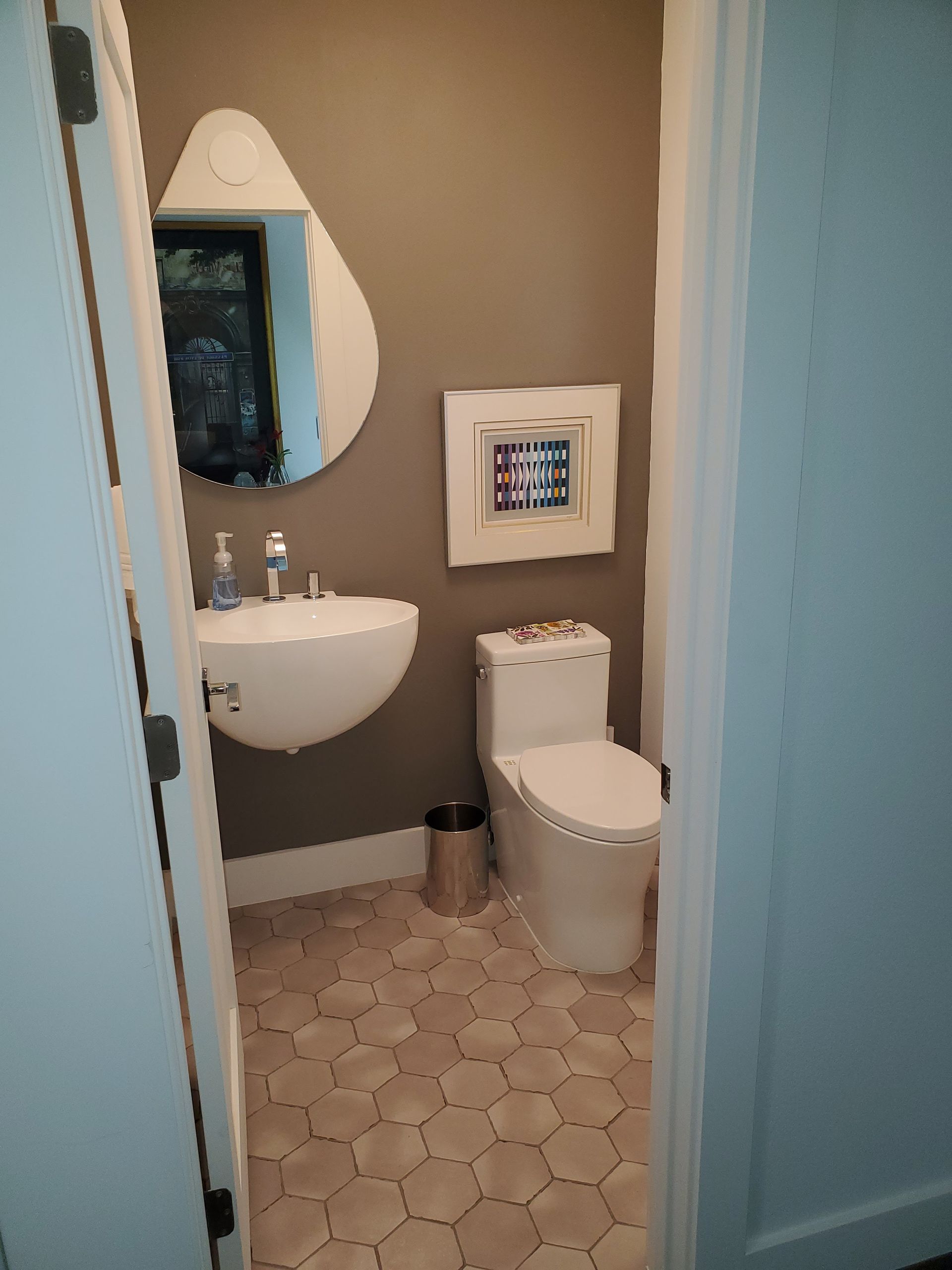 A small powder room with a white wall-mounted sink, a toilet, a teardrop-shaped mirror, and hexagon floor tiles.