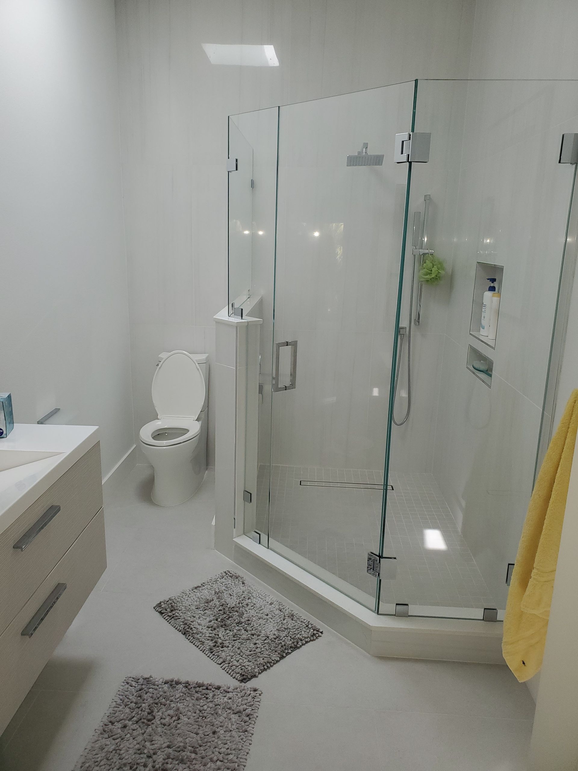 A modern bathroom with a light wood vanity, a toilet, a glass-enclosed corner shower, and two grey speckled bath mats.