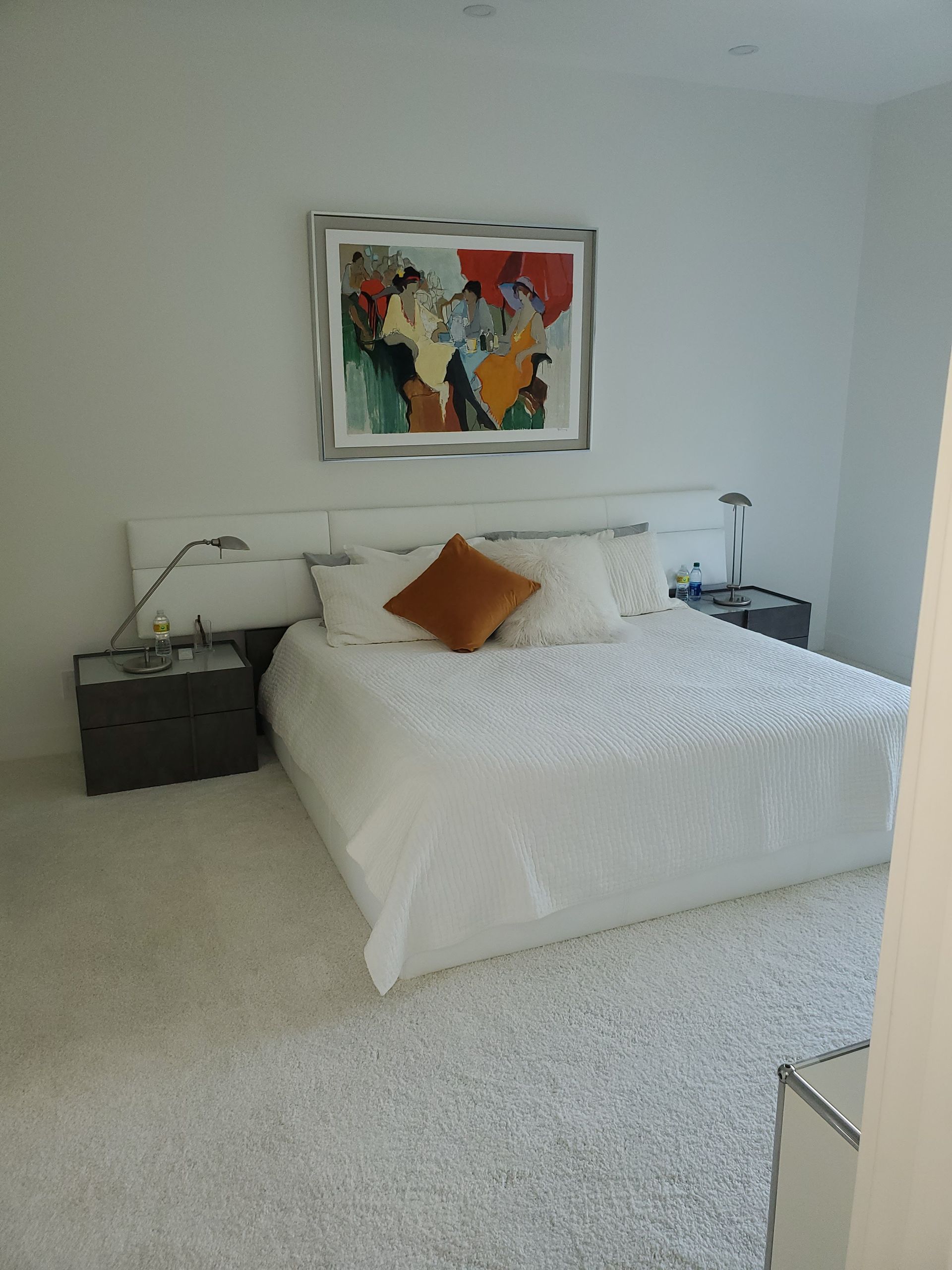 A modern bedroom with a white bed, a vibrant abstract painting above the headboard, and bedside tables on a white carpet.