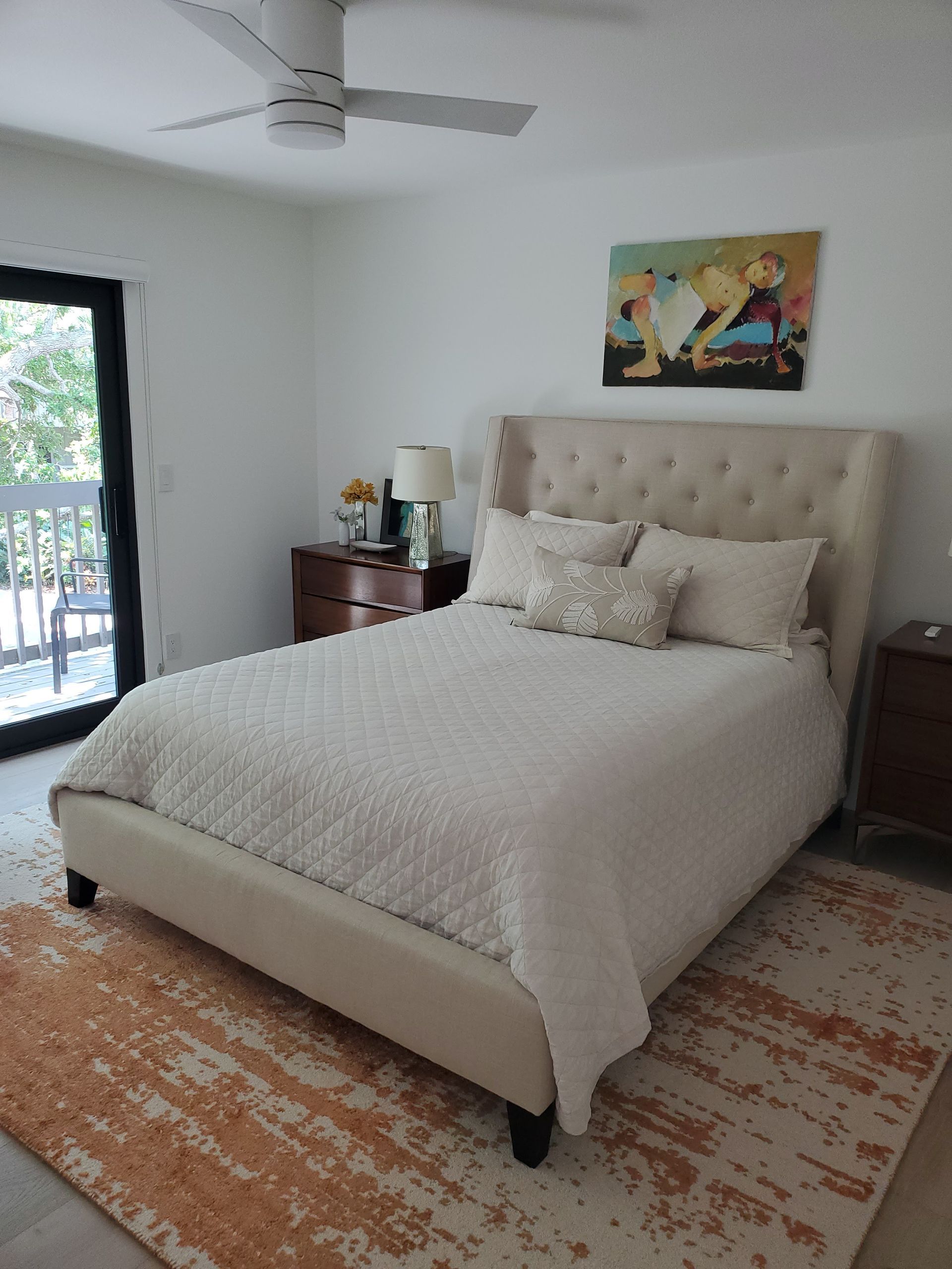 A bedroom with a tufted beige bed, cream bedding, an orange patterned rug, and a painting above the headboard.