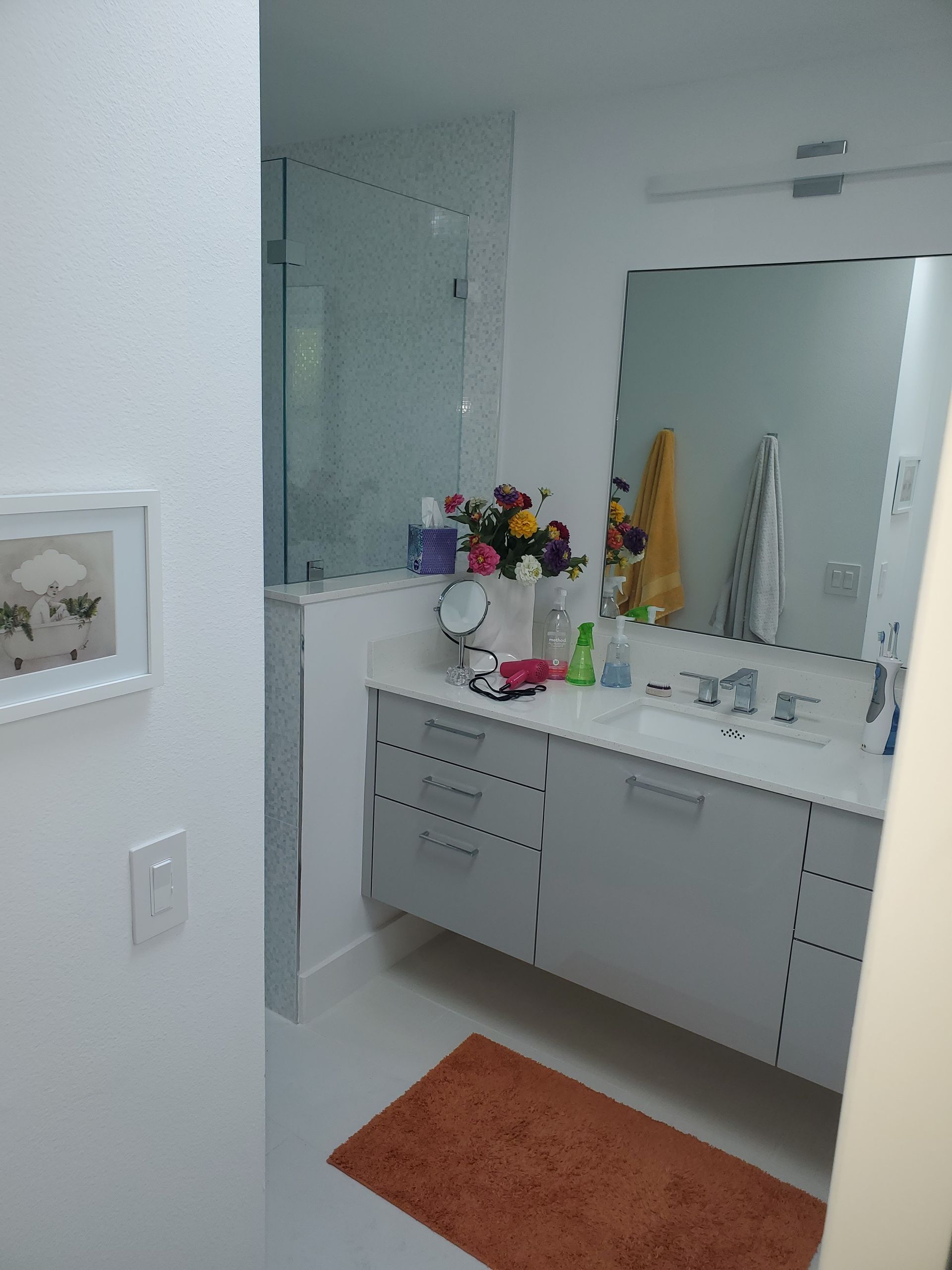 A modern bathroom with light gray cabinetry, a rectangular mirror, a glass-enclosed shower, and an orange bath mat.