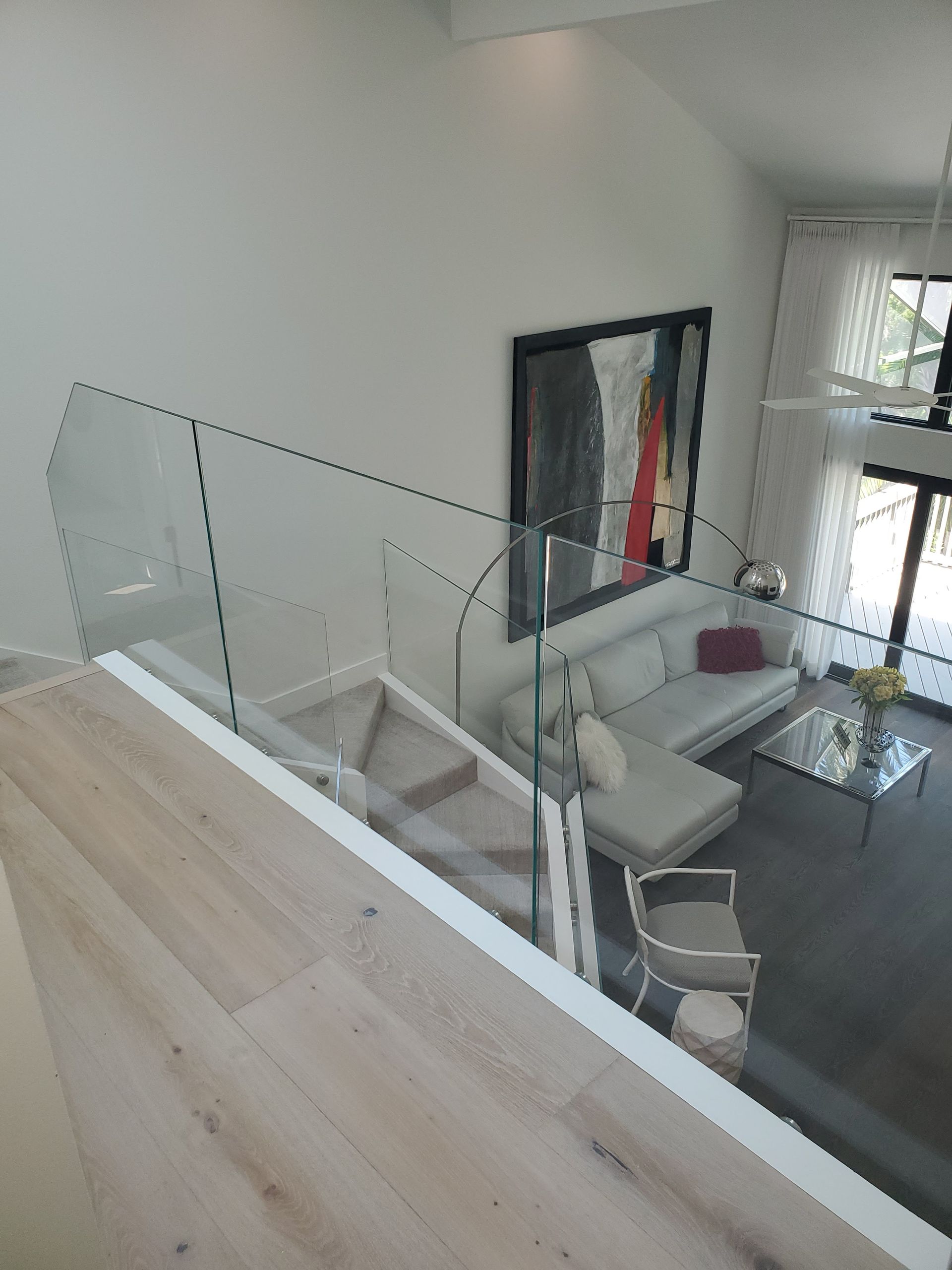 A high-angle view looking down from a landing with glass railings onto a modern living room with a white sofa and art.