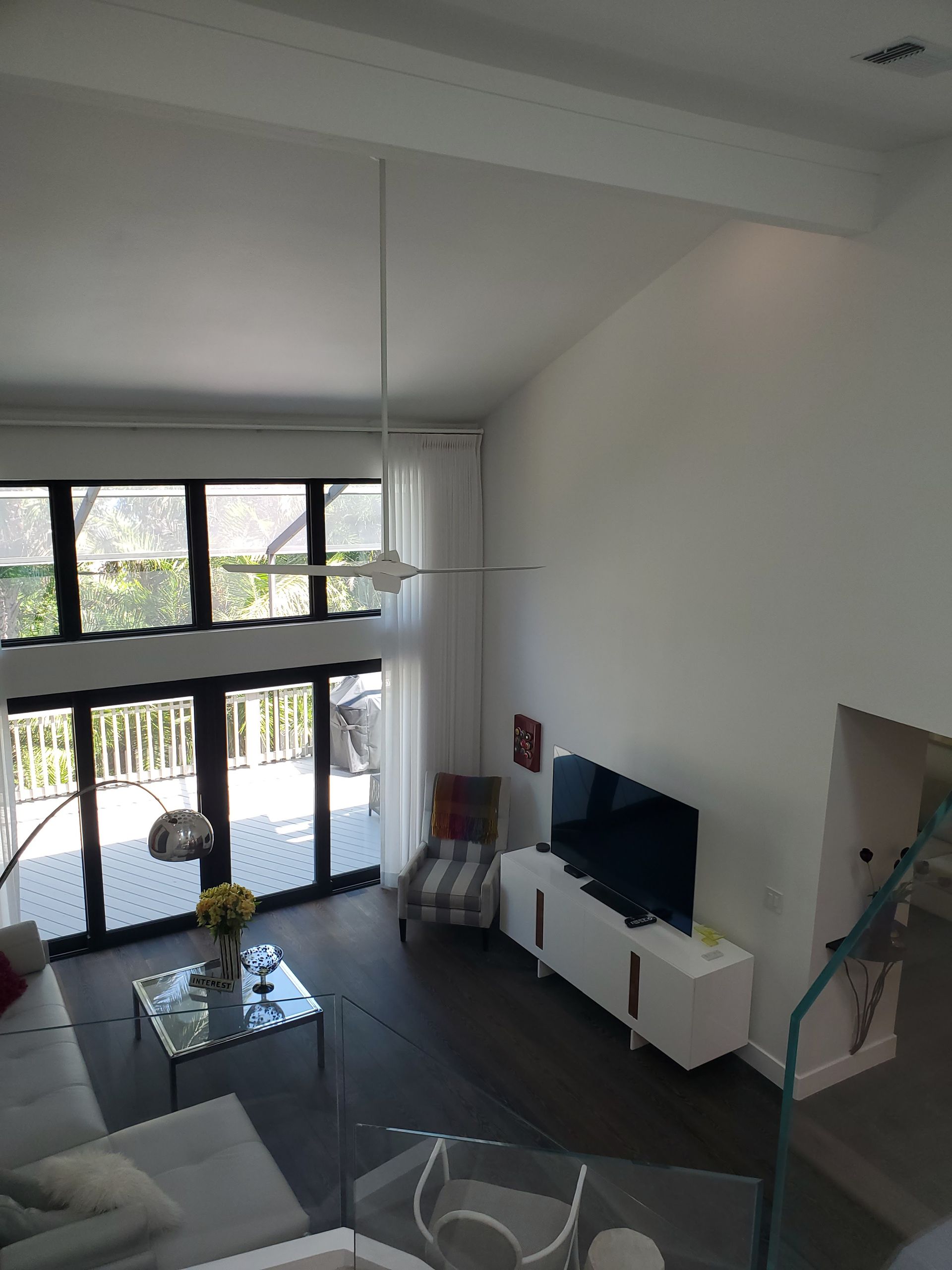 A high-angle view of a living room with dark floors, large floor-to-ceiling windows, white walls, and a television console.