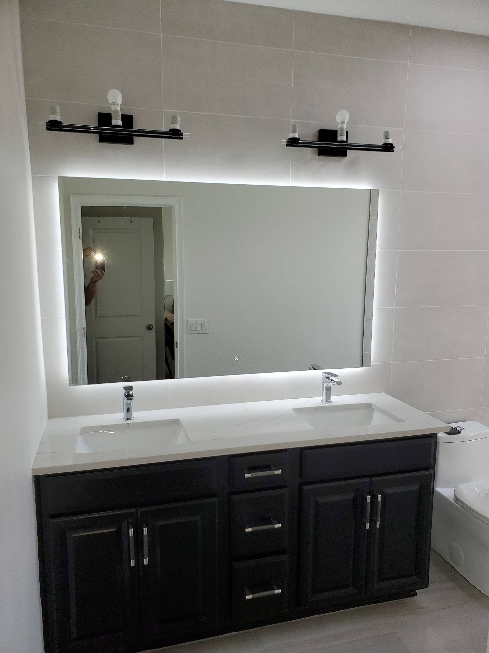 A modern bathroom vanity with a dark wood cabinet, white countertop, two sinks, a backlit rectangular mirror, and sconces.