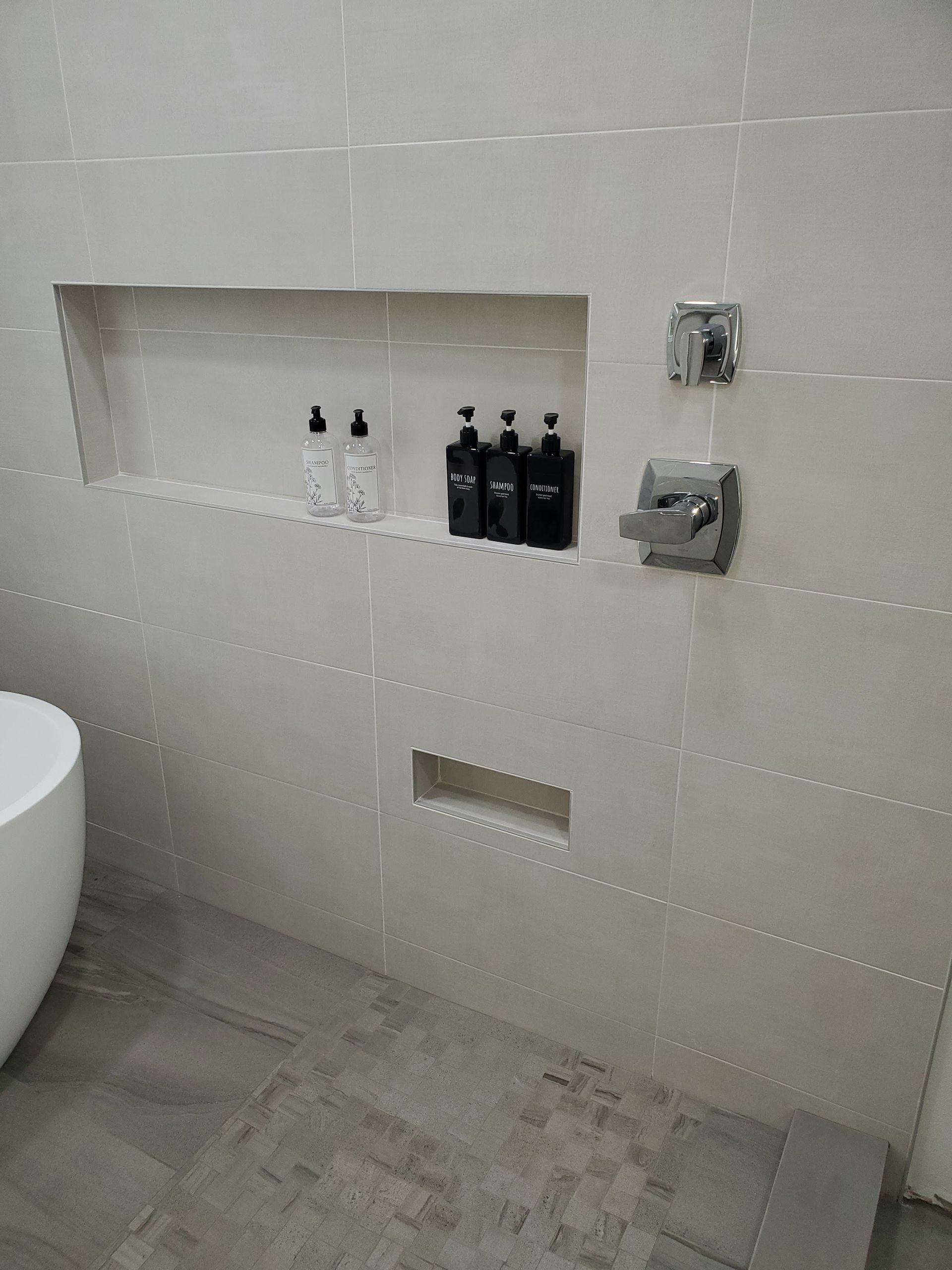 A tiled shower wall with a recessed shelf holding soap bottles, a smaller niche below, and wall-mounted chrome fixtures.
