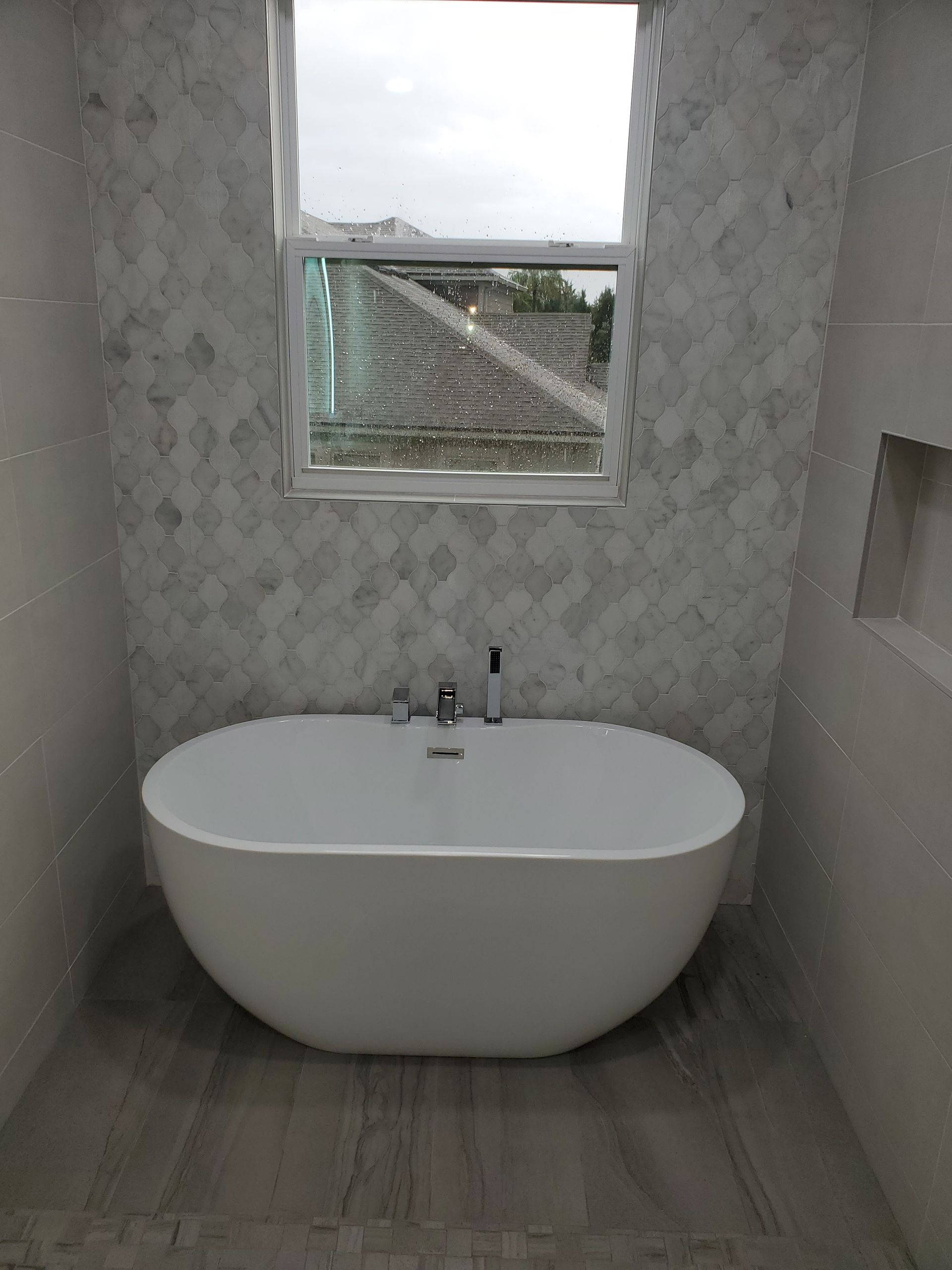 A modern white freestanding bathtub centered in a tiled bathroom alcove beneath a window.