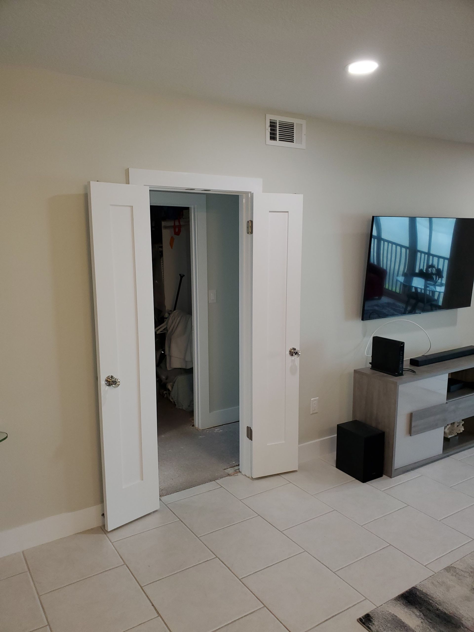 An open white double door leads into a closet next to a wall-mounted television and a grey media console on tile flooring.