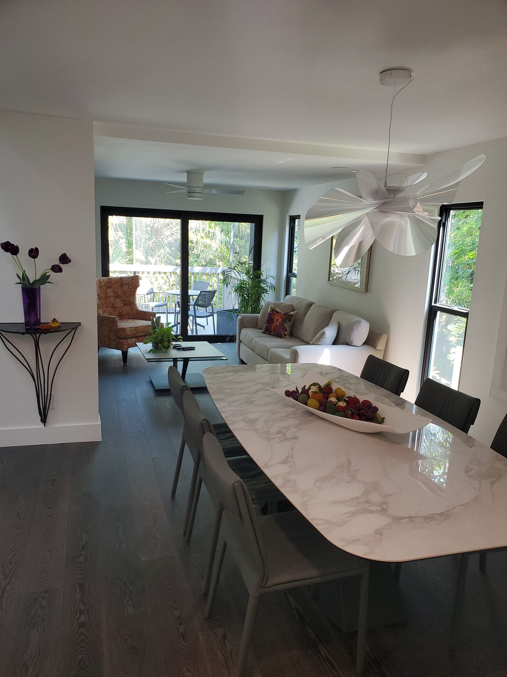 Modern dining room with a white marble table and gray chairs, opening into a sunlit living area with a patio view.