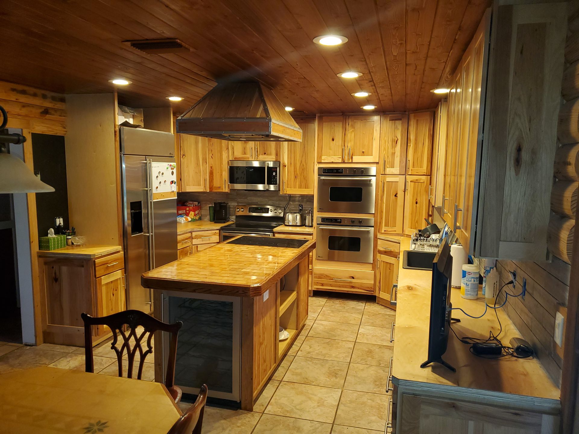 A rustic kitchen with wood-paneled walls and ceiling, a wooden island, stainless steel appliances, and tiled floors.