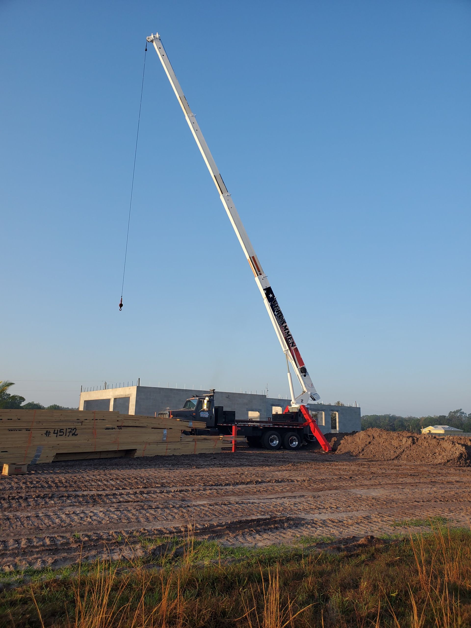 A tall crane with a white boom stands on a dirt construction site against a clear blue sky.