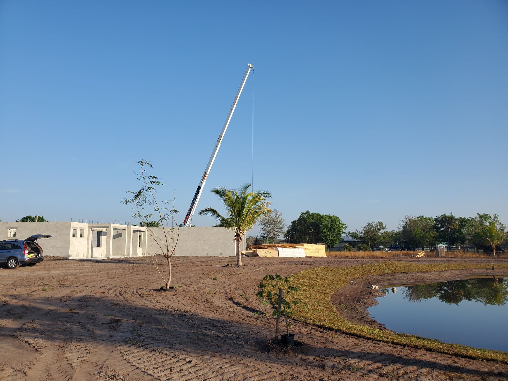 A crane stands near a construction site featuring a small building, trees, and a pond under a clear blue sky.