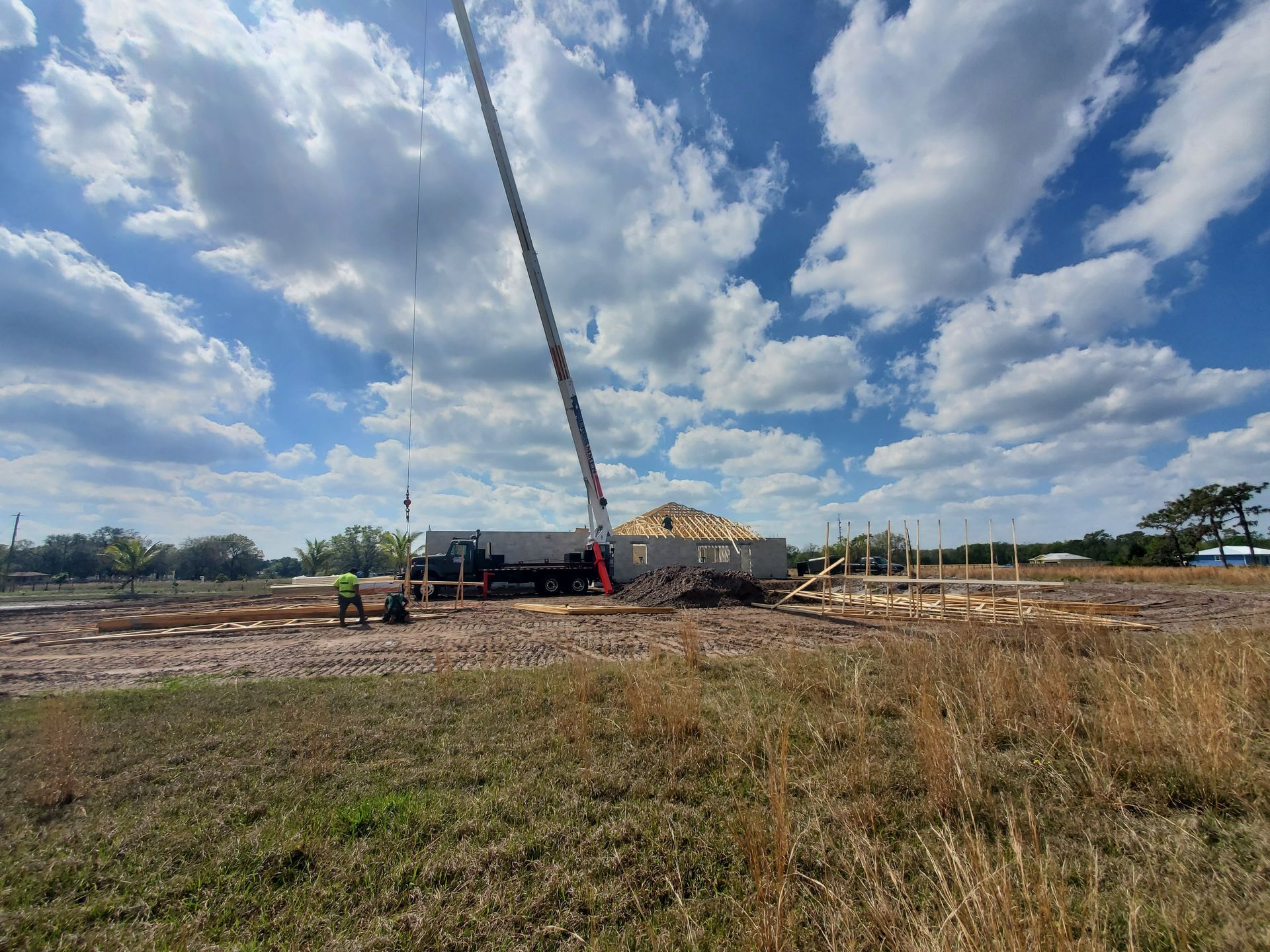 A construction site with a crane lifting roof trusses over a house frame under a bright, cloudy sky.