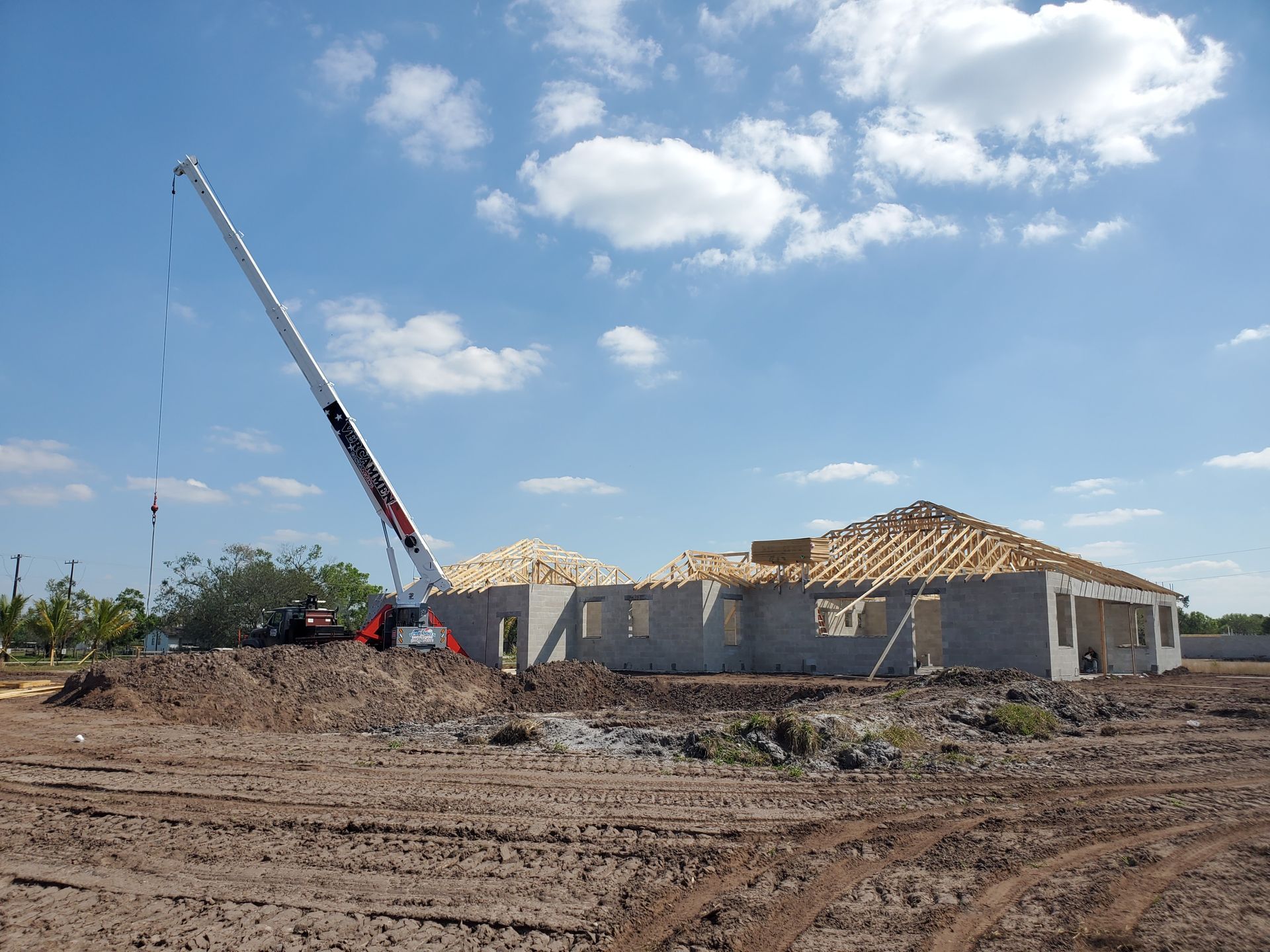 A construction crane stands beside a house under construction with a wooden roof frame on a dirt lot under a blue sky.
