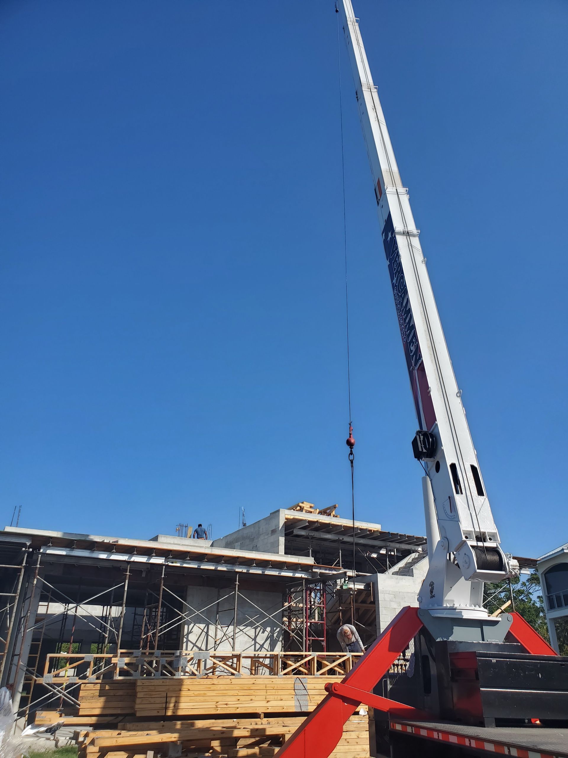 A white crane with an extended boom stands against a clear blue sky at a construction site with wood scaffolding.
