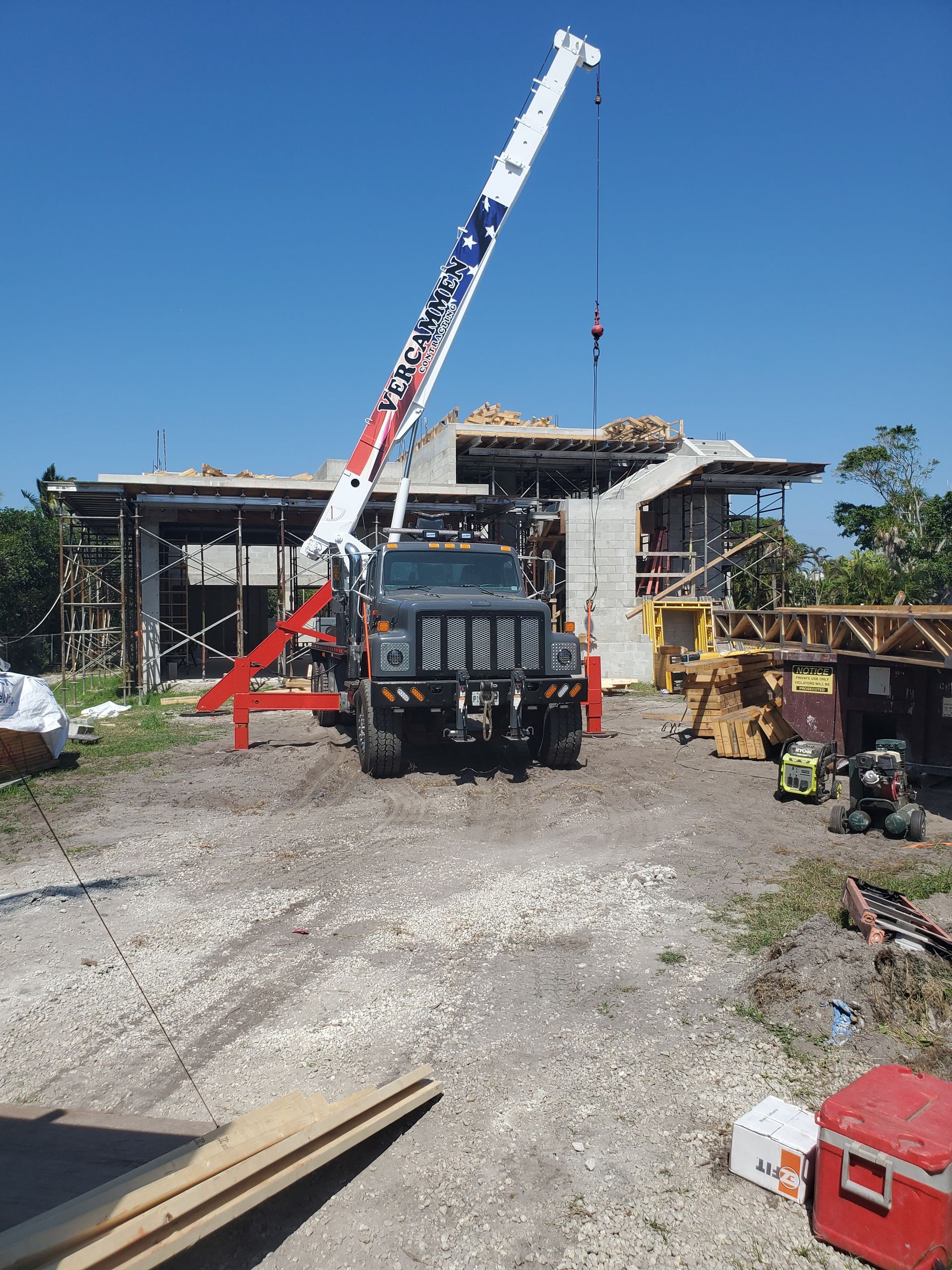 A crane truck with a raised boom positioned in front of a construction site with building materials on a sunny day.