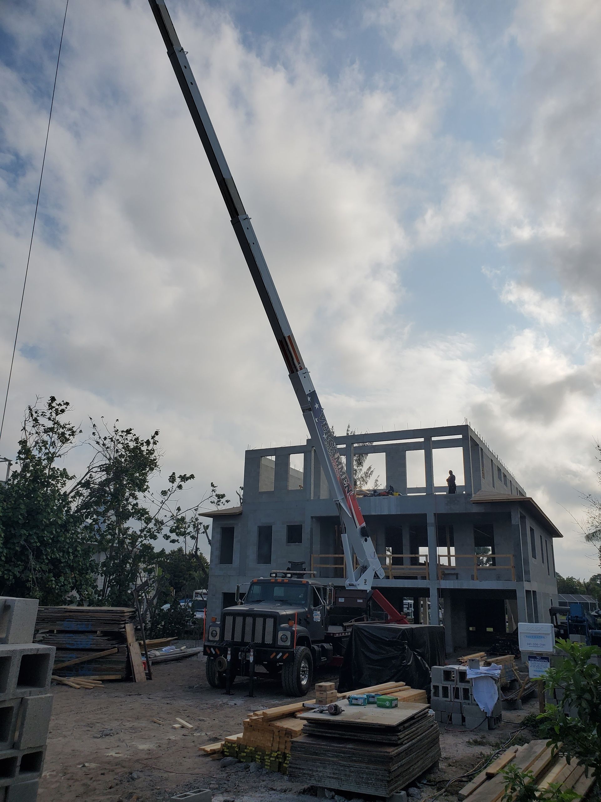 A crane lifts construction materials toward the upper level of a gray, multi-story building under a cloudy sky.