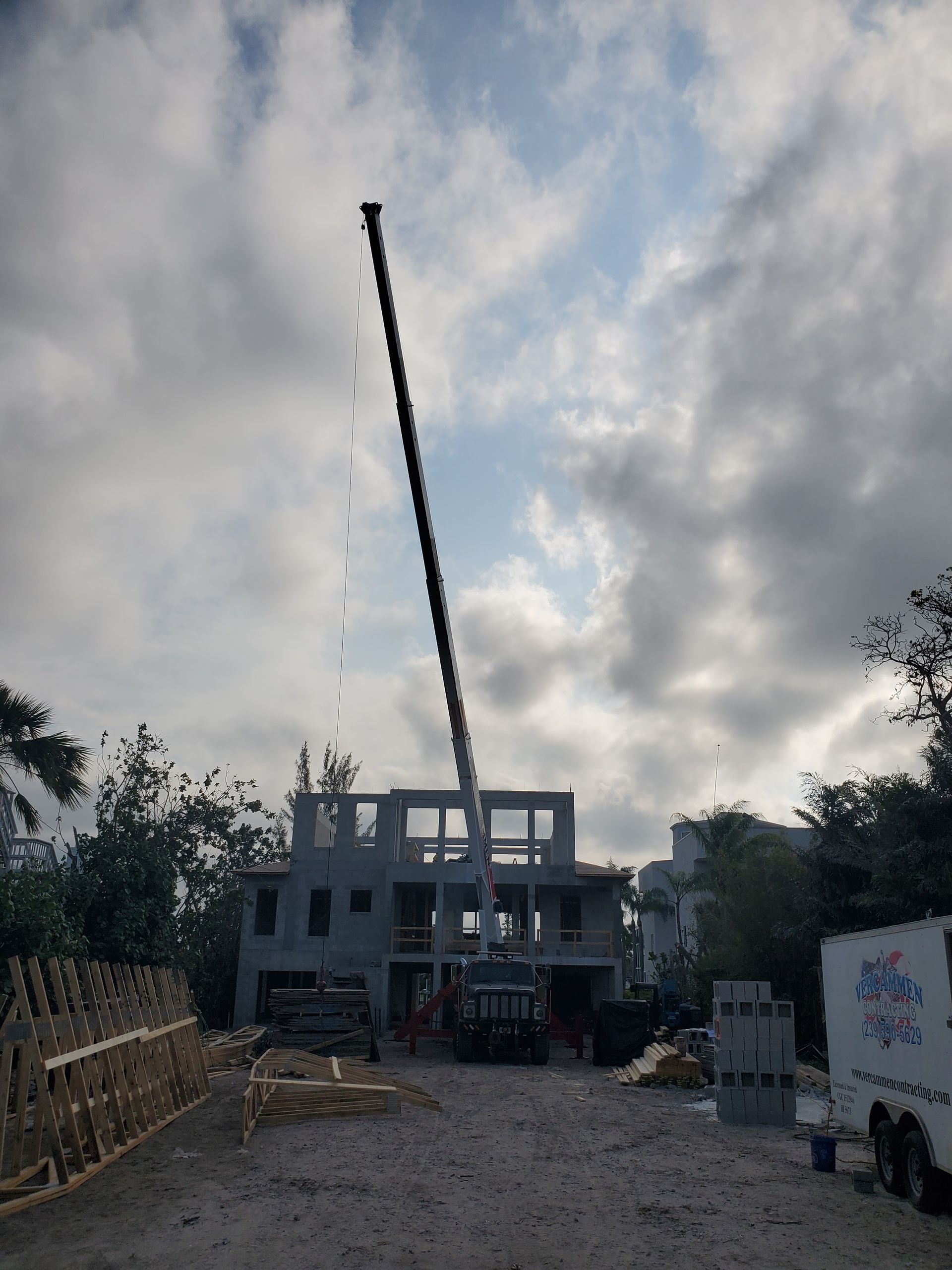 A construction crane with an extended boom stands in front of a multi-story concrete building under a cloudy sky.