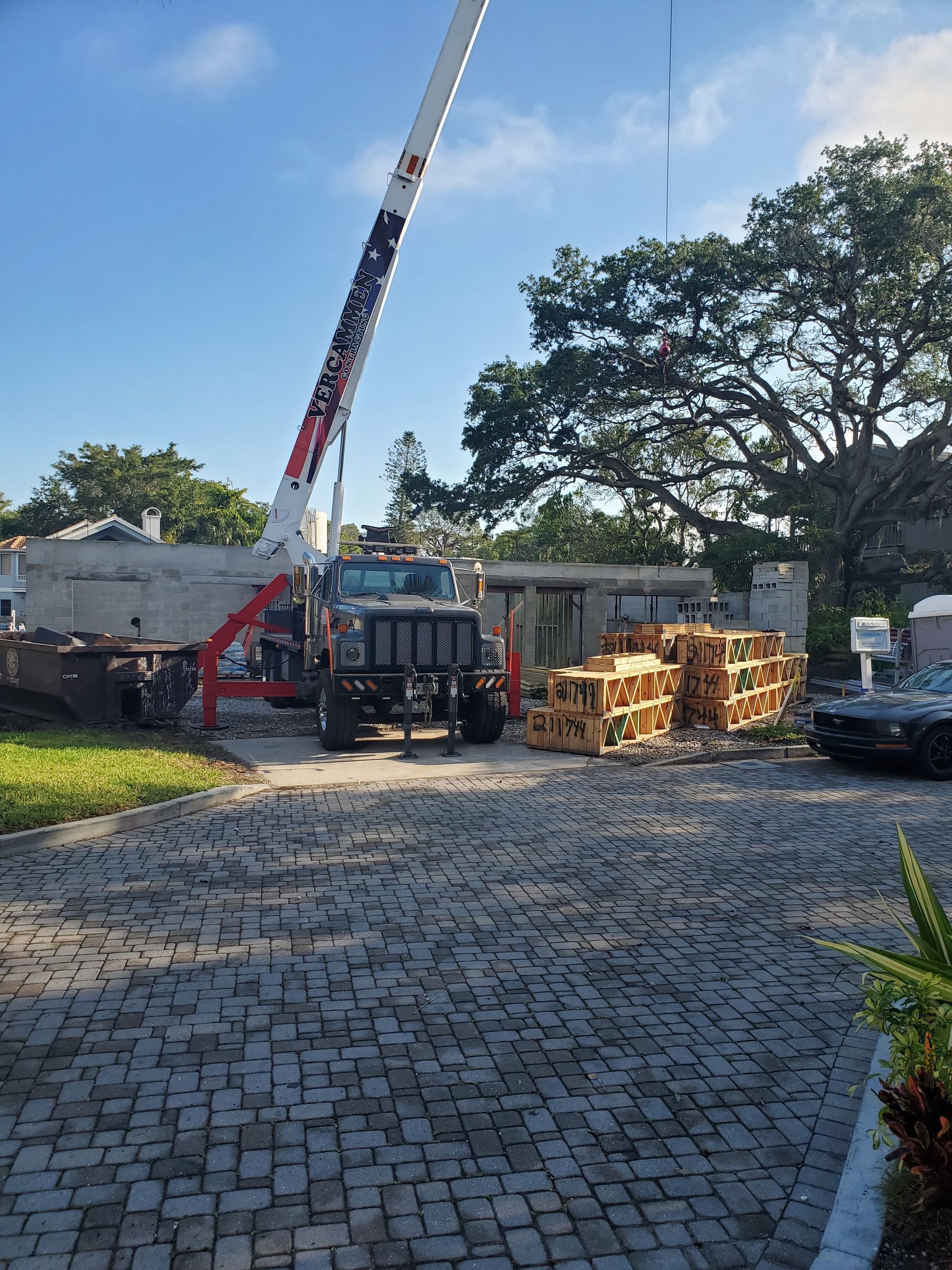 A construction crane stands in a driveway next to a partially demolished house and stacked pallets on a sunny day.