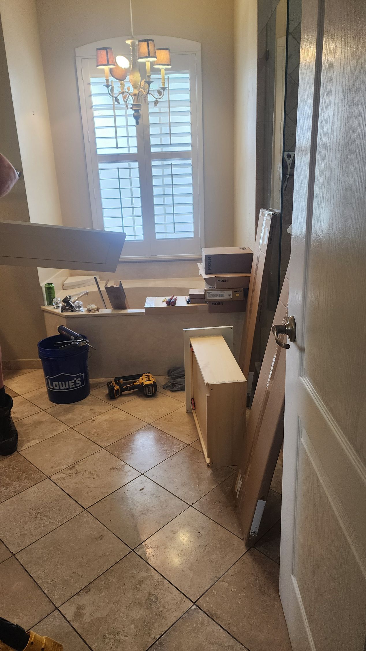 A messy bathroom under renovation with light-colored tile, a bathtub, stacked furniture components, and a chandelier.