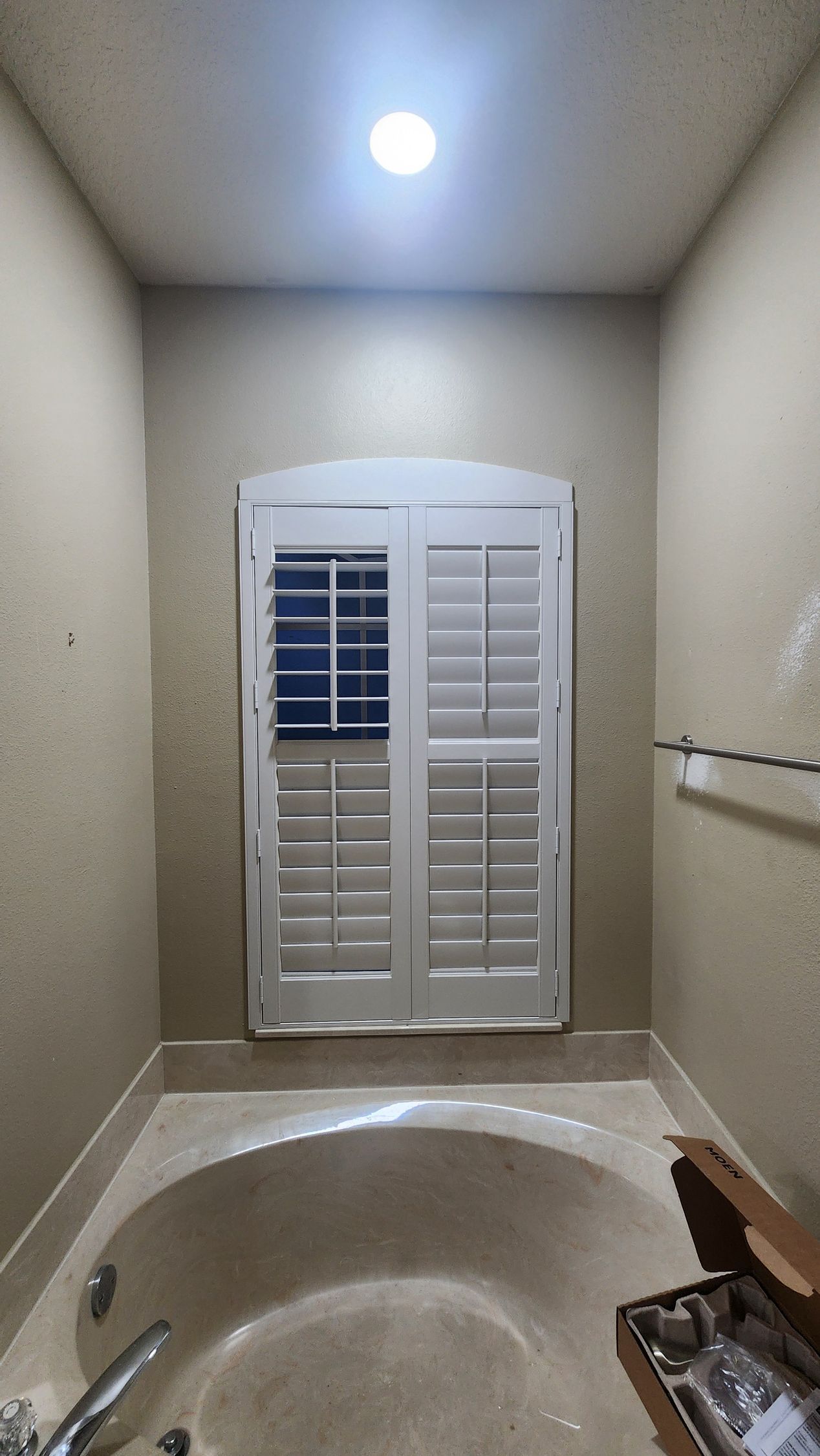 A white plantation shutter installed on an arched window above a round bathtub in a tan-walled bathroom.