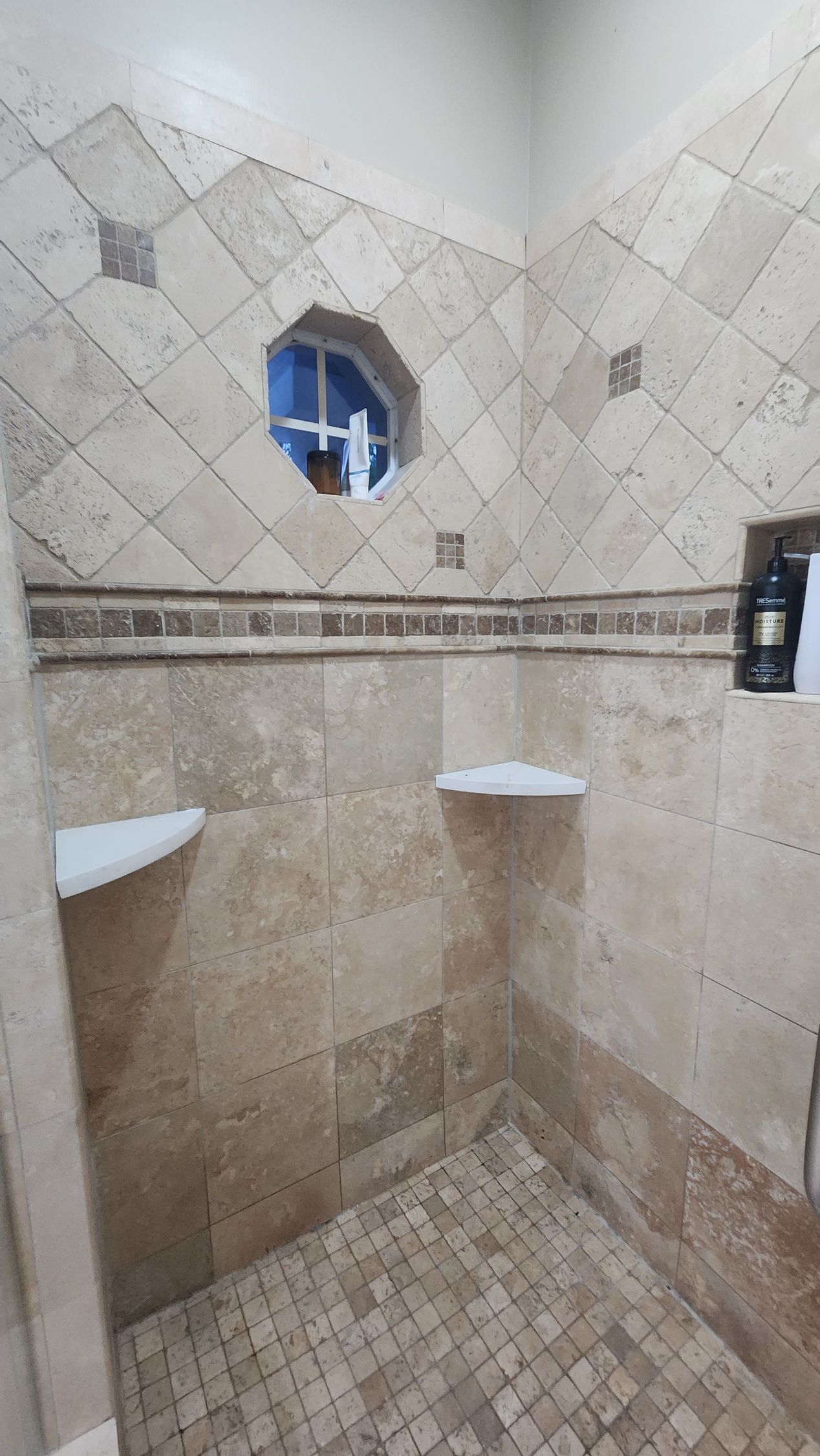 A shower with beige natural stone tile walls, a decorative border, two corner shelves, and an octagonal window.