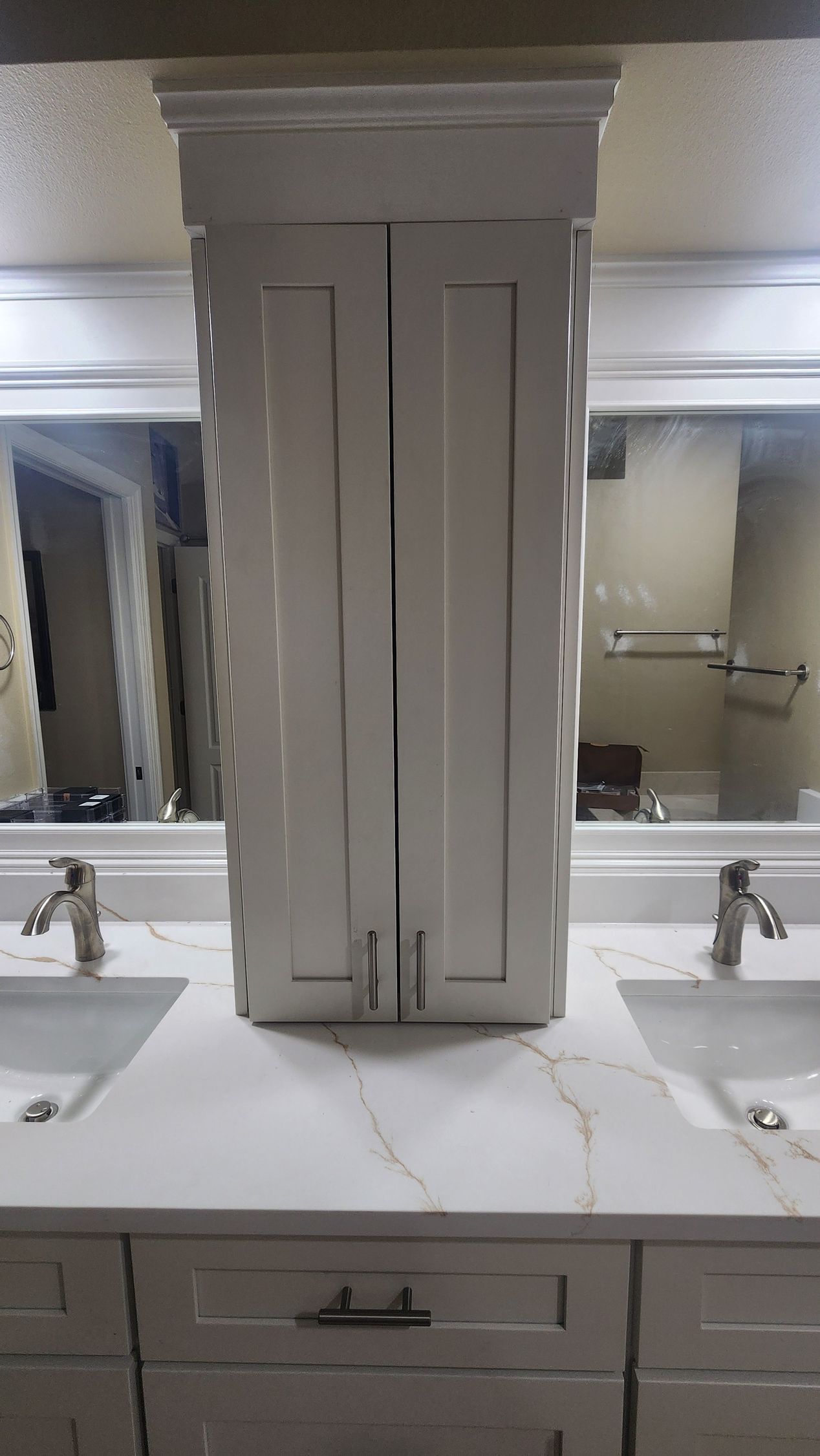 A double-sink bathroom vanity with a central, white cabinet tower between two mirrors, featuring white marble countertops.