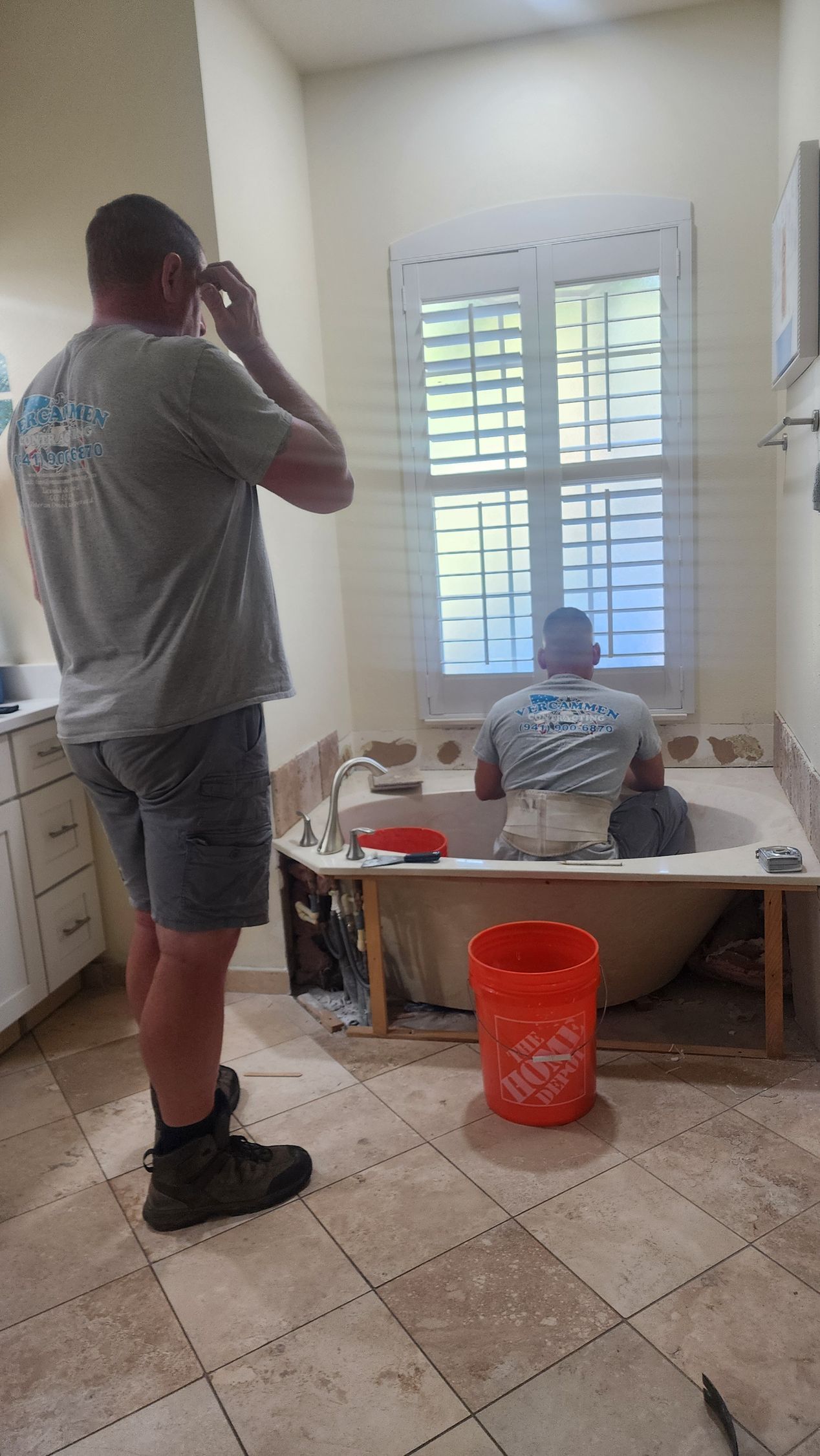 Two individuals in work uniforms renovating a bathroom, one standing and one sitting inside a large bathtub.