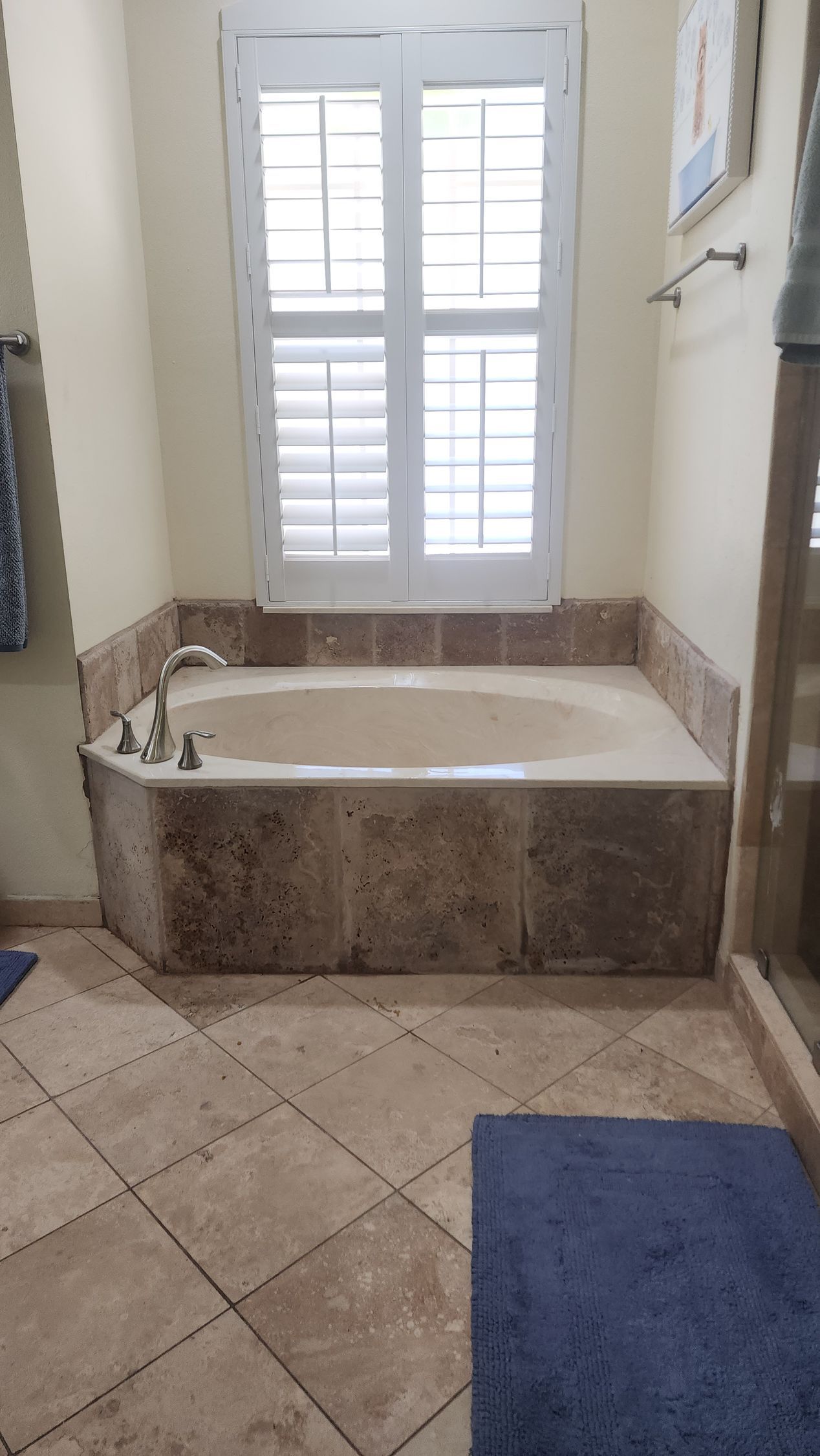 A built-in soaking tub with beige stone tile surround set beneath a shuttered window in a bathroom.