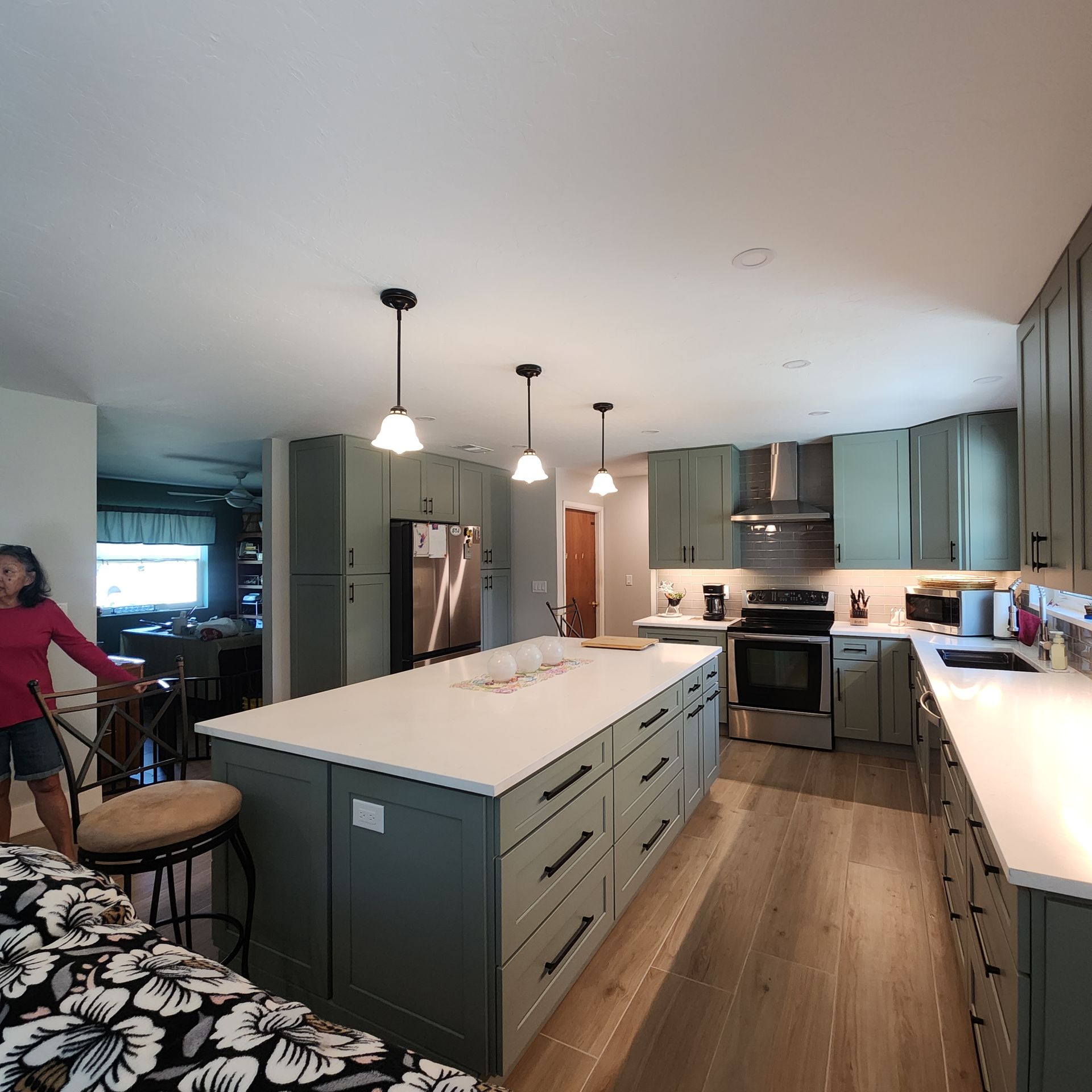 A bright, modern kitchen features dark green cabinets, a large white-topped island, three pendant lights, and wood floors.