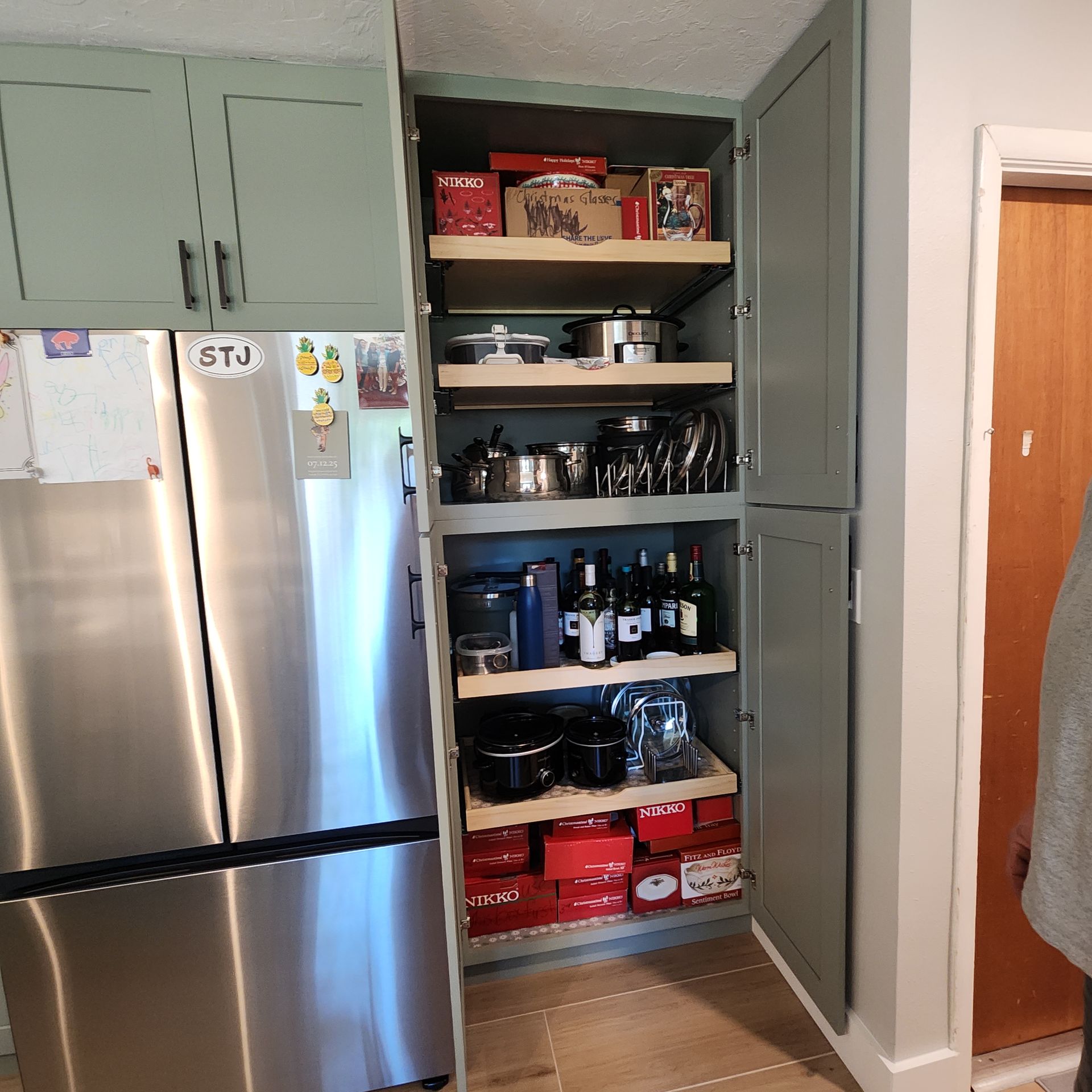 A tall, sage green pantry cabinet with open slide-out shelves stands next to a stainless steel refrigerator.
