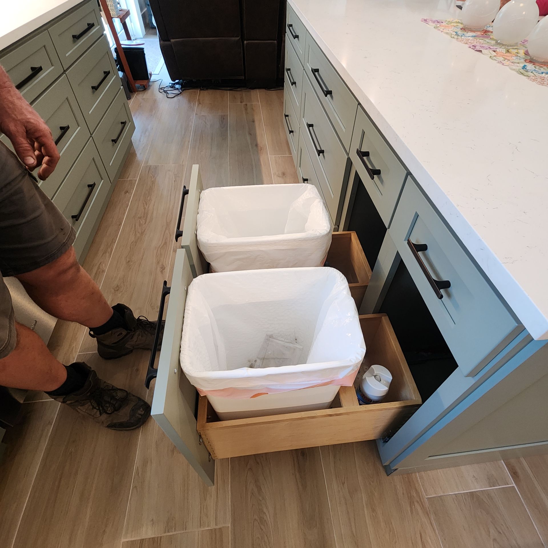 A person stands in a kitchen next to an open pull-out drawer containing two white trash bins.