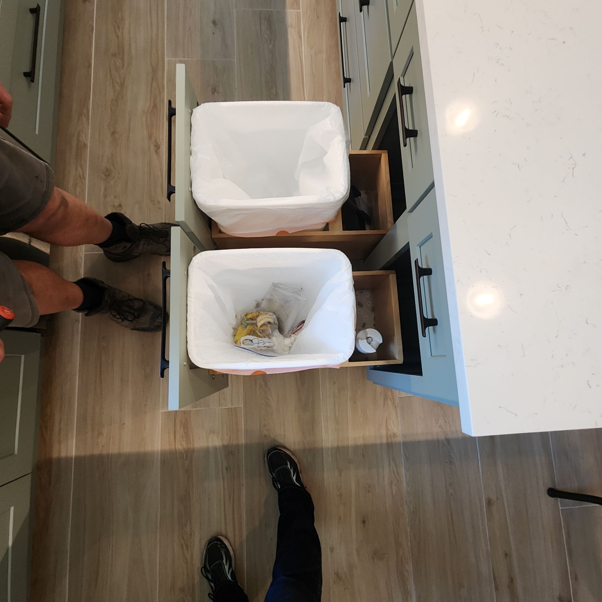 An overhead view of a kitchen island pull-out drawer containing two trash bins, with legs and feet visible on the floor.