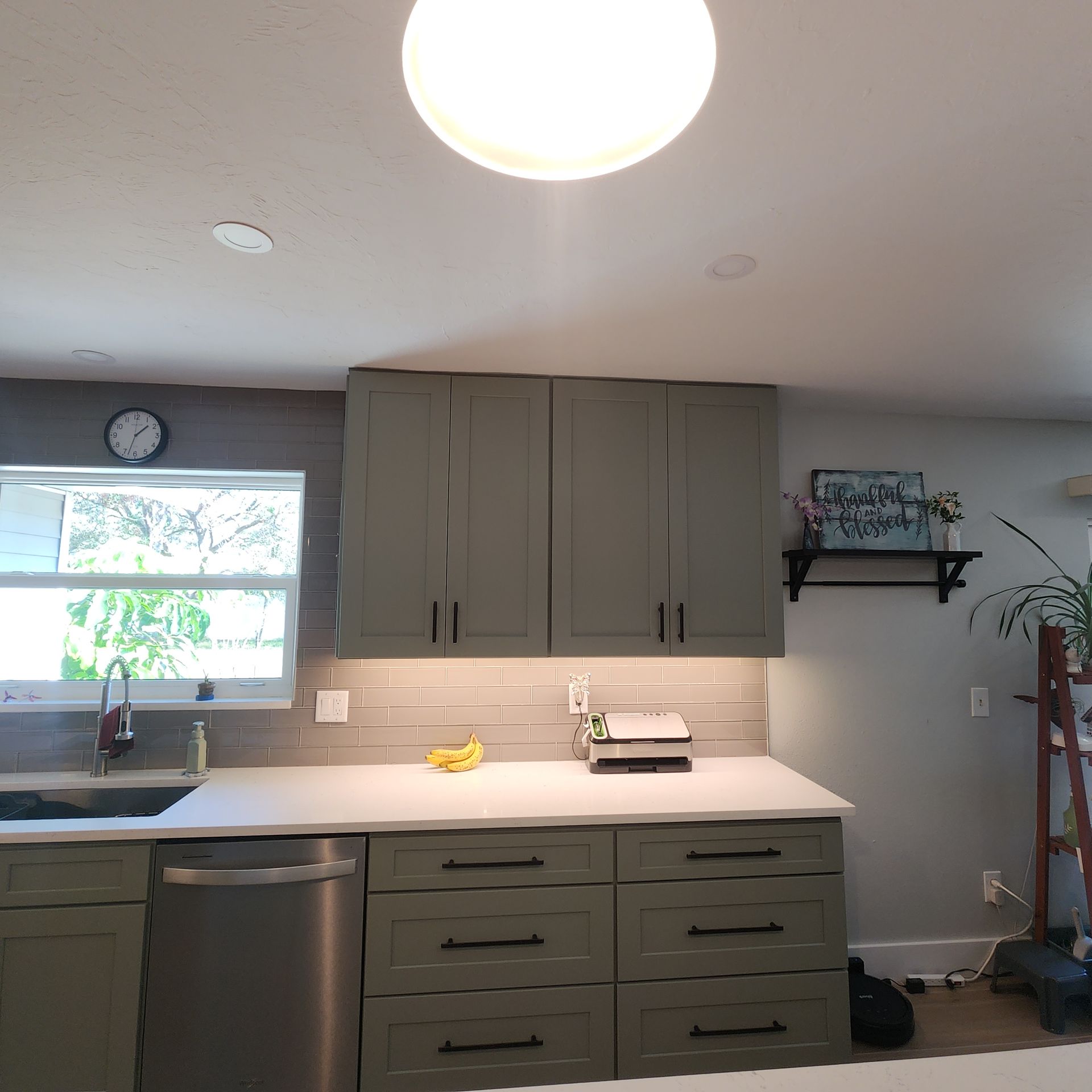 A kitchen with sage green cabinets, a gray tiled backsplash, a white countertop, and an under-cabinet light.