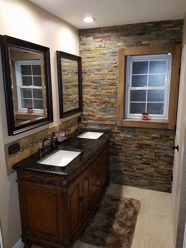 A bathroom with a double-sink vanity, stone accent wall, rustic wood-framed window, and two dark-framed mirrors.
