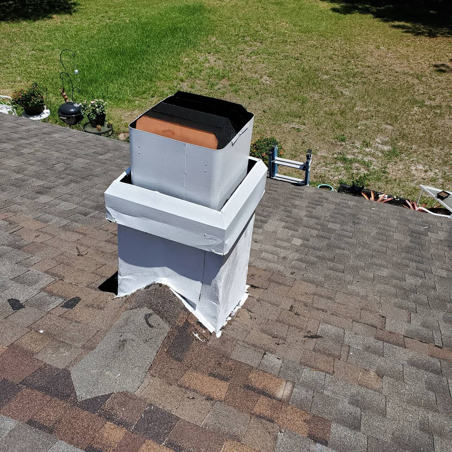 A white chimney structure with a square metal top and inner flue, positioned on a sloped, shingled roof outdoors.
