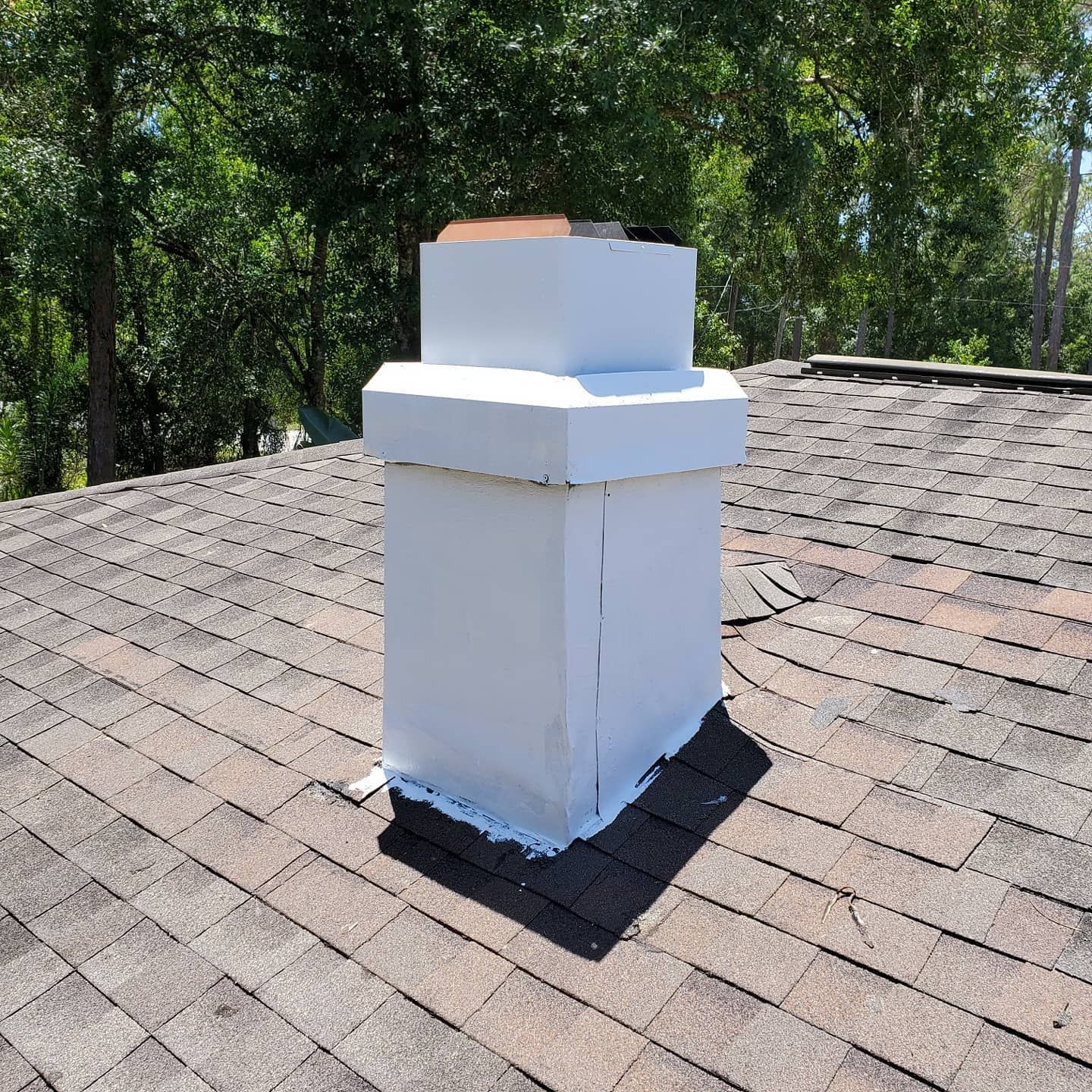 A white-painted rectangular chimney sits on a shingled roof surrounded by trees under a clear blue sky.