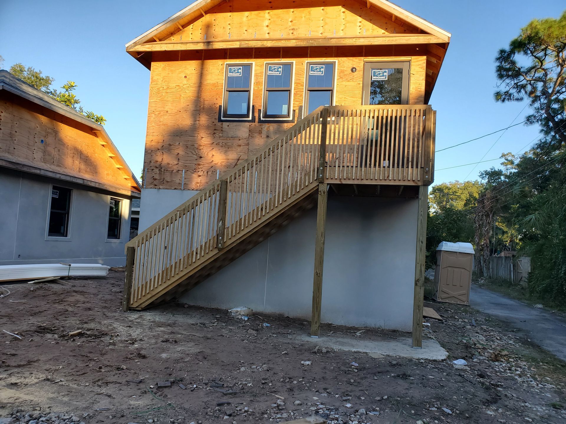 An unfinished two-story building under construction with a wooden exterior deck and stairs, situated on a dirt lot.