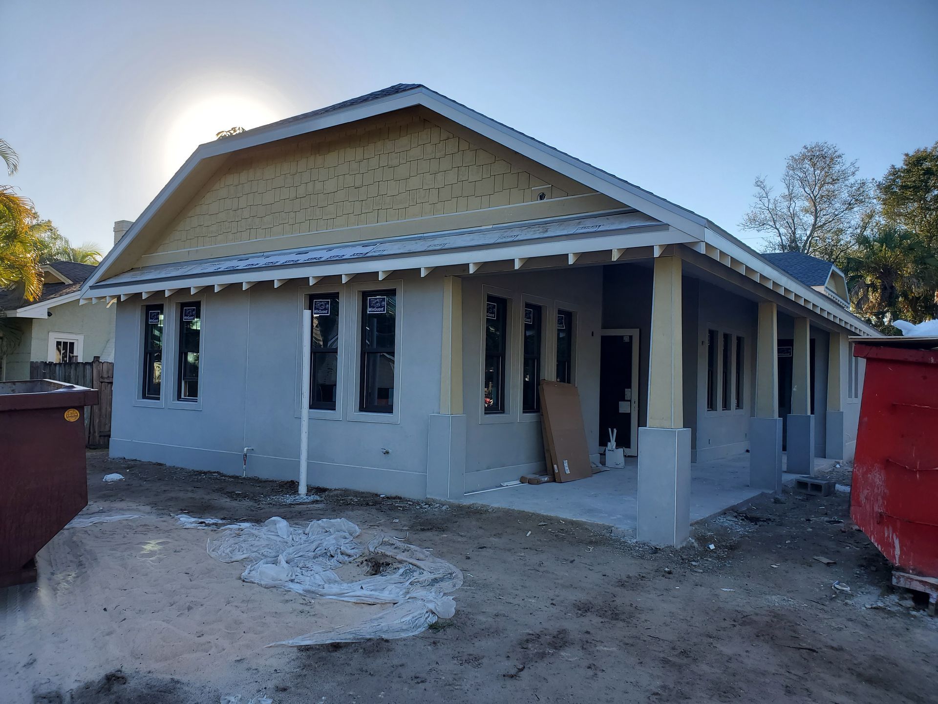 A single-story house under construction with light gray walls, a yellow gable end, and a wrap-around porch with pillars.