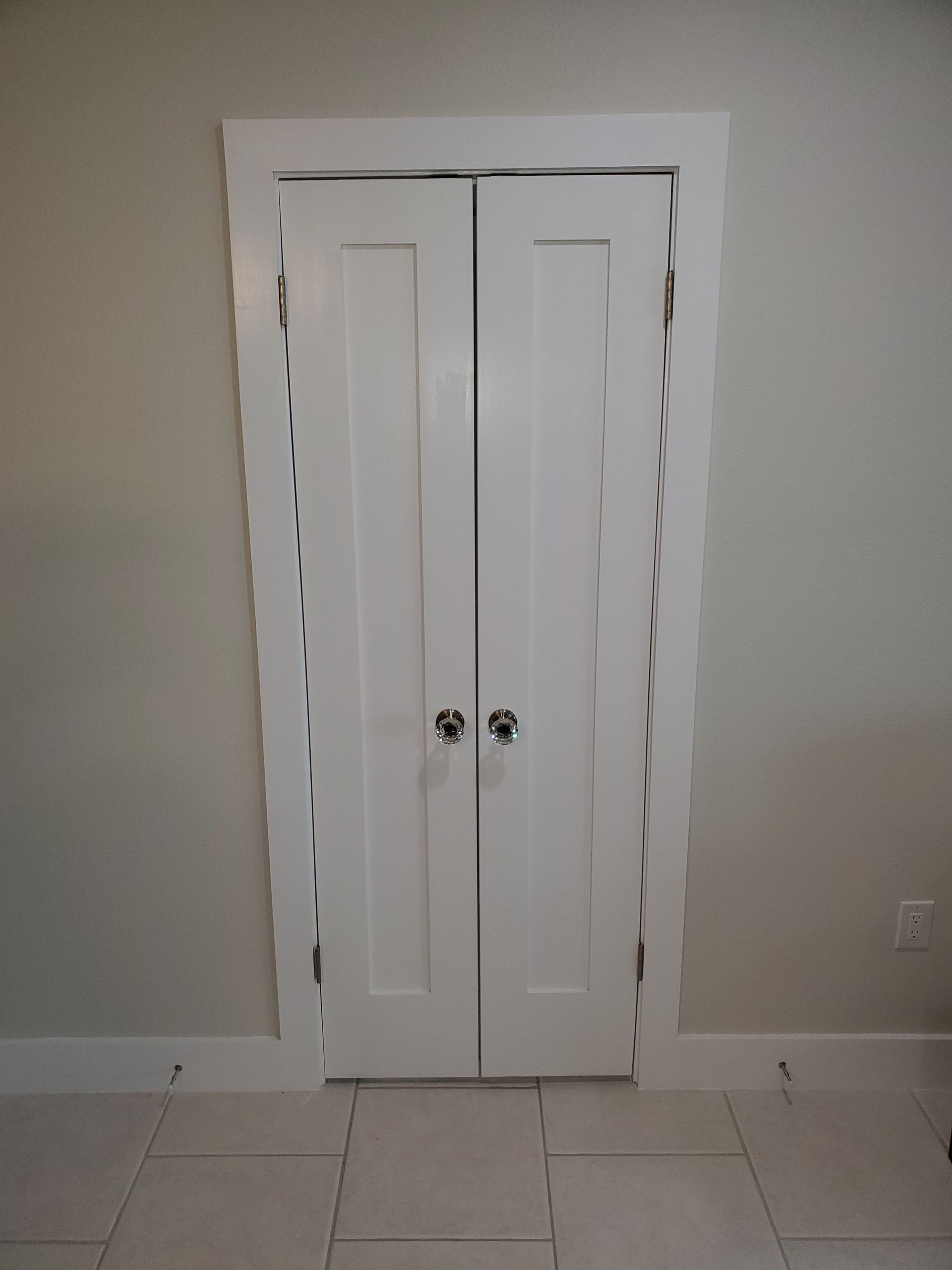 White double closet doors with central knobs, installed in a light-colored wall with white door trim and baseboards.