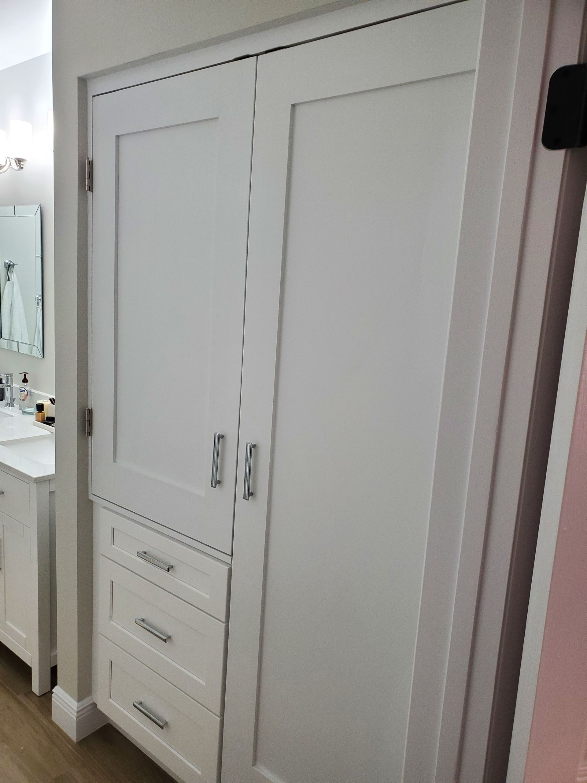 A white floor-to-ceiling cabinet with three lower drawers and two tall doors, situated next to a bathroom vanity.